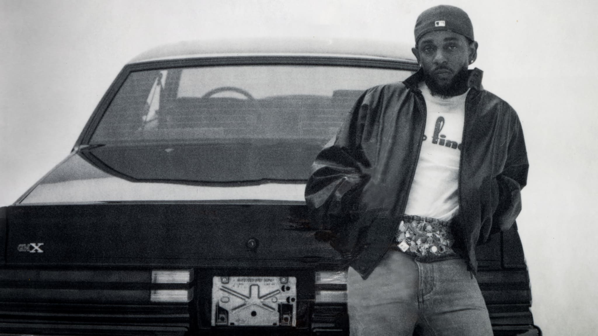 Kendrick in a casual outfit leans against the back of a car, wearing a cap, jacket, and jeans, with a confident expression.