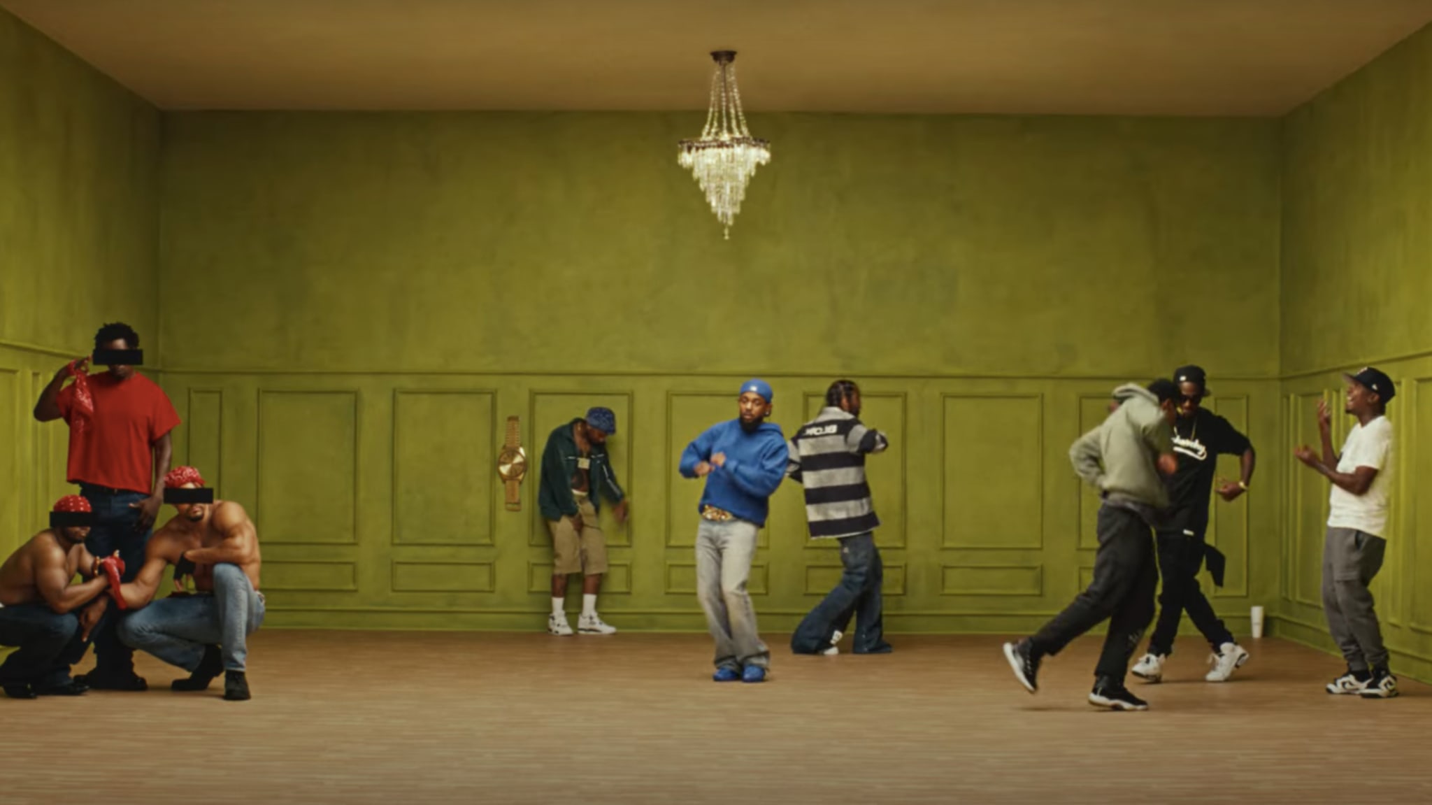 A group of people in a green room, some dancing and others standing or crouching. A chandelier hangs from the ceiling.