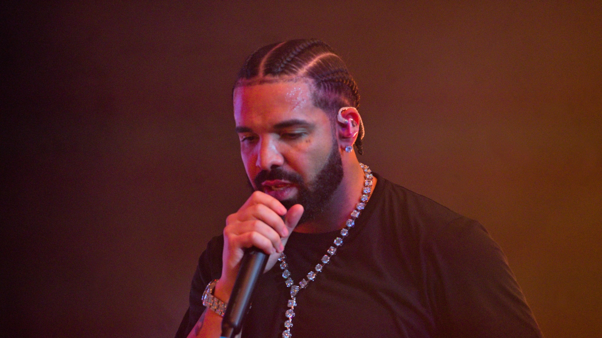 Drake performing on stage, wearing a black shirt and a diamond necklace, holding a microphone.
