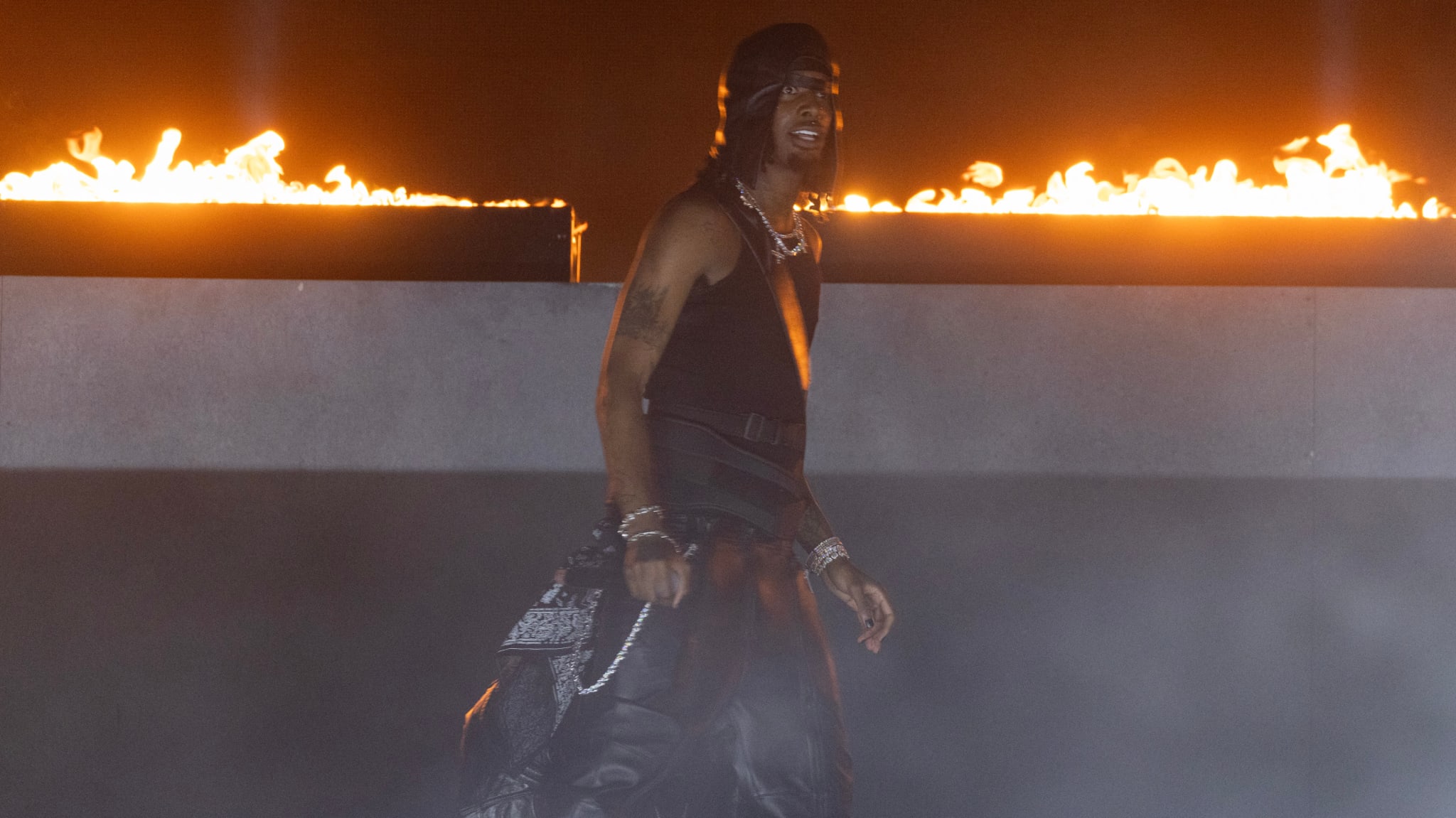 Carti in a black outfit and cap walks in front of a fiery background, exuding a dramatic and intense atmosphere.