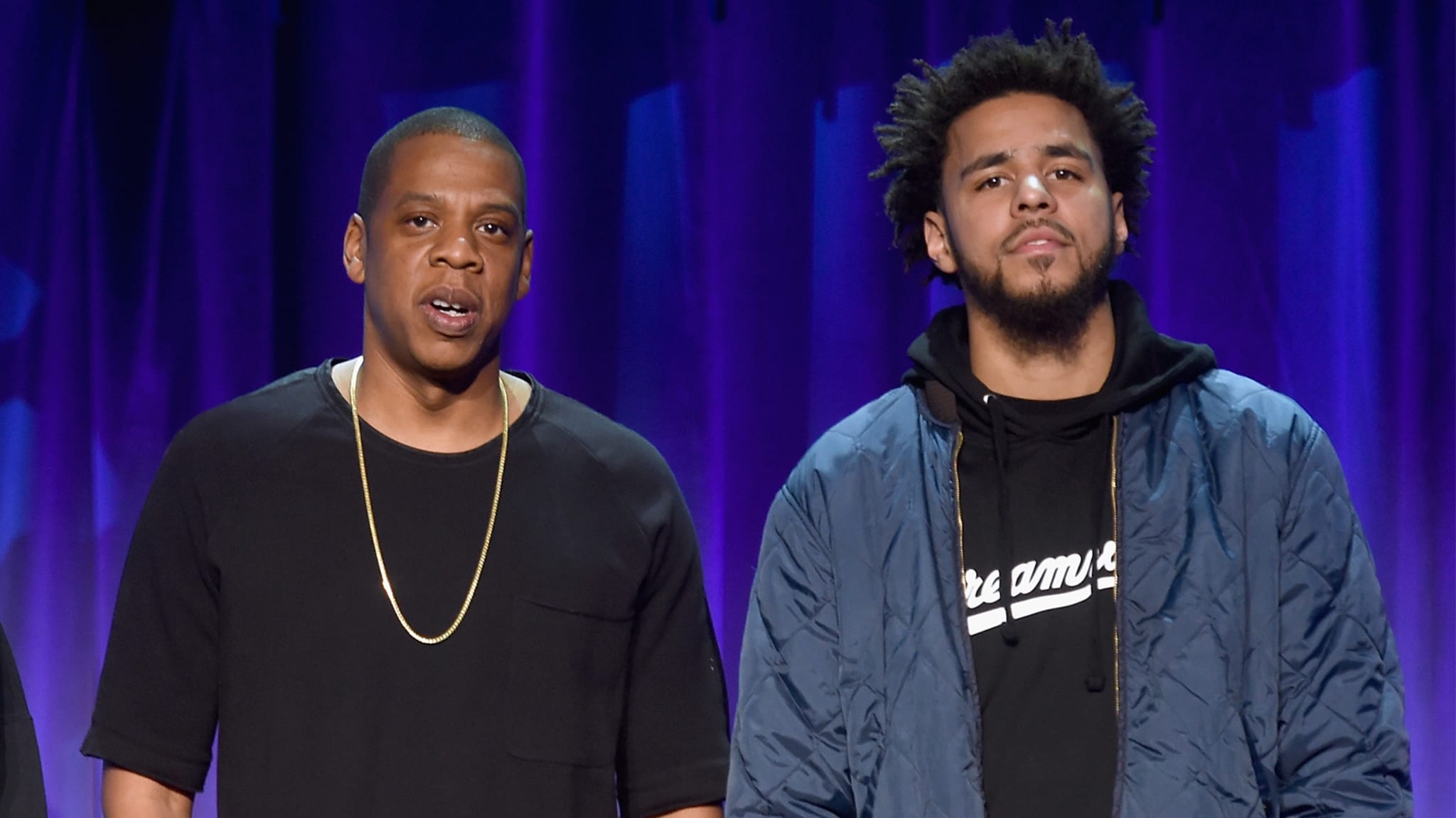 Jay-Z and J. Cole standing together on stage, with Jay-Z in a black shirt and J. Cole in a blue jacket over a hoodie.