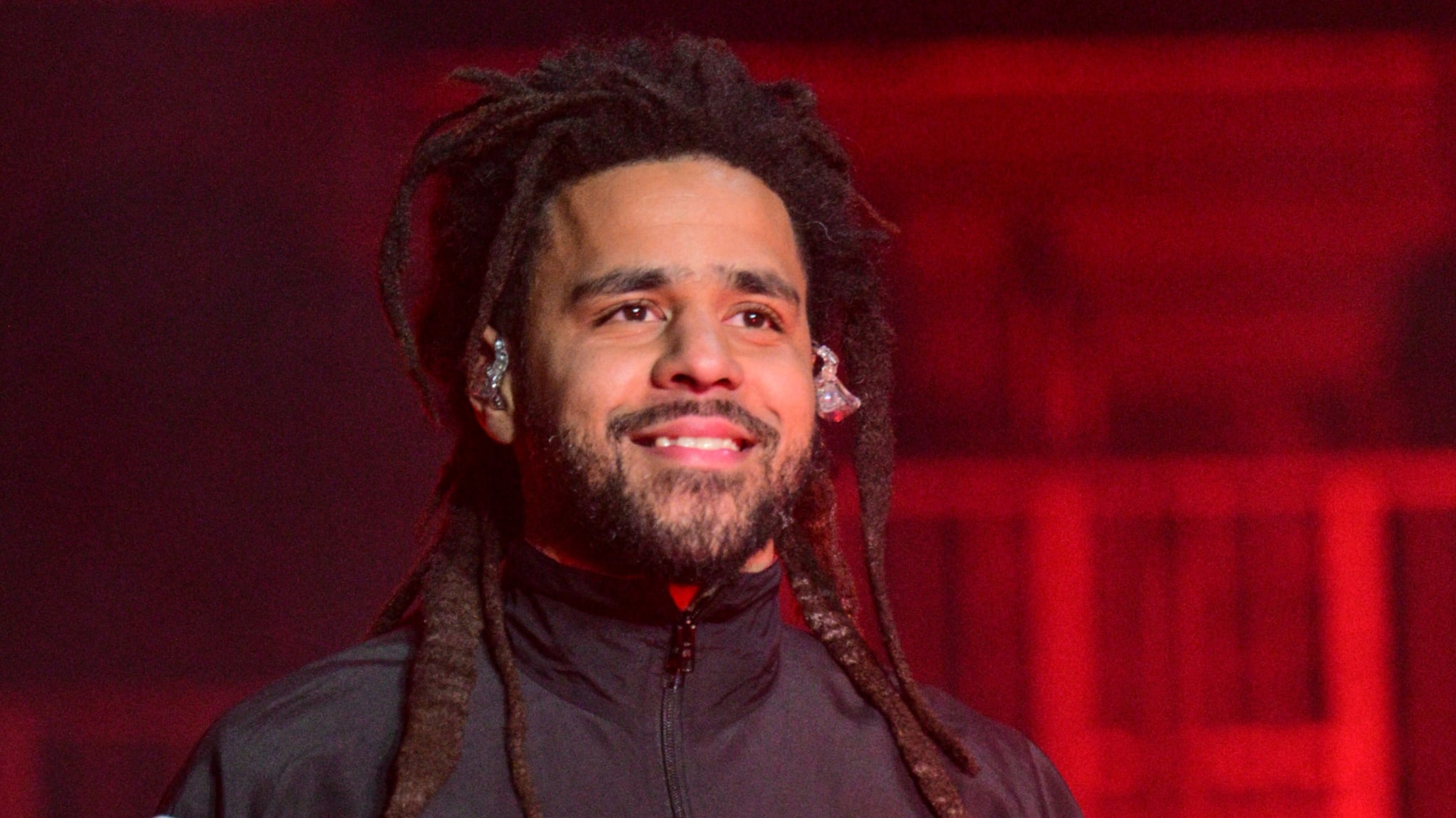 J. Cole, wearing a black jacket, smiles on stage with red lighting in the background.