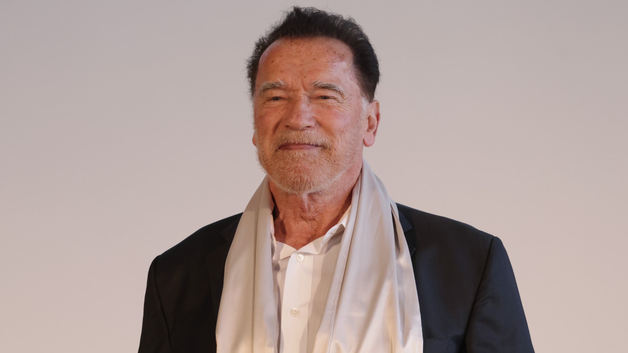 Arnold Schwarzenegger, wearing a suit and scarf, smiles against a plain background.