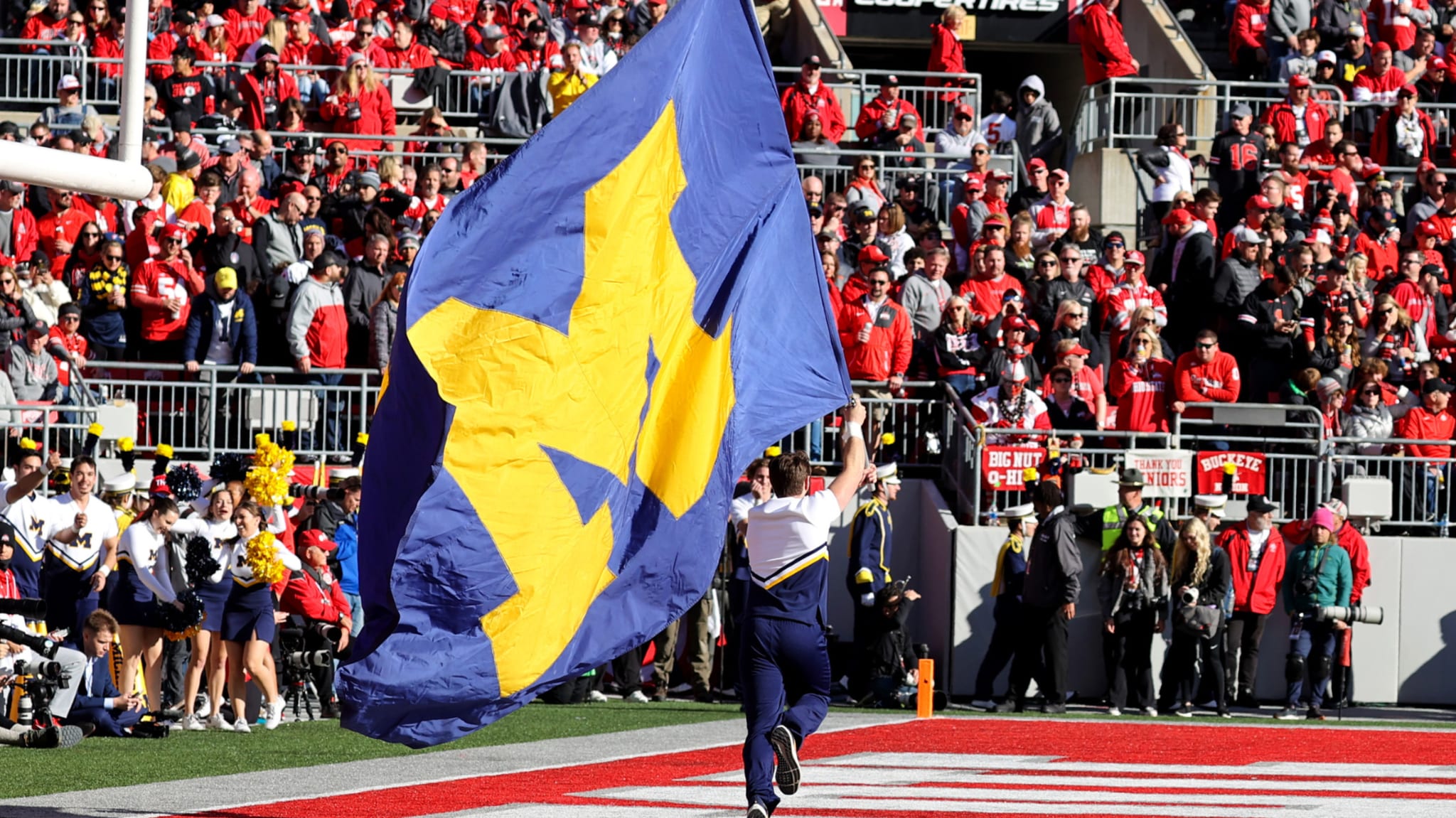 A cheerleader runs across a football field with a large University of Michigan flag. A crowd in red fills the stadium seats.