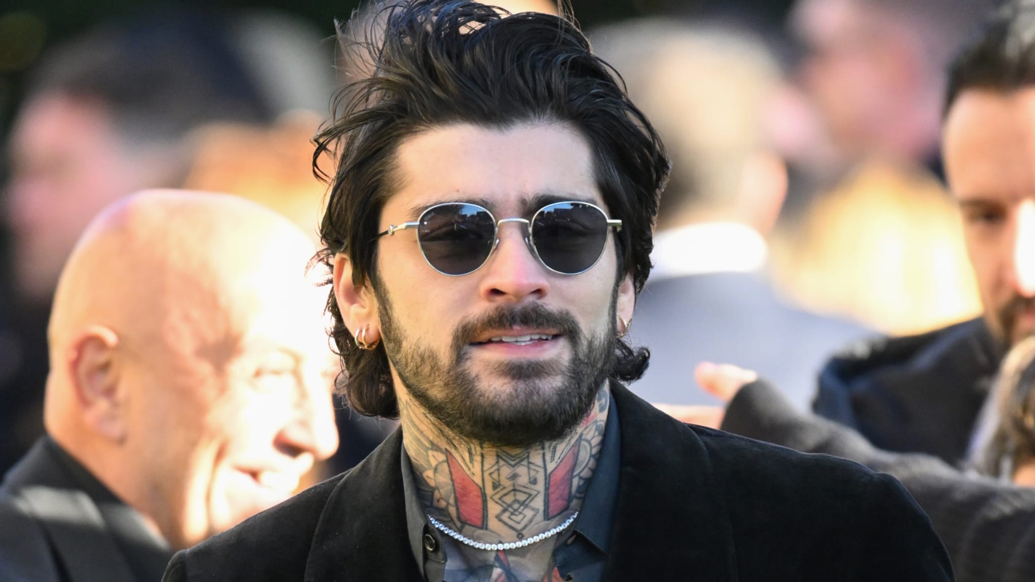 A man with dark hair, sunglasses, and neck tattoos, wearing a black jacket in a crowd.
