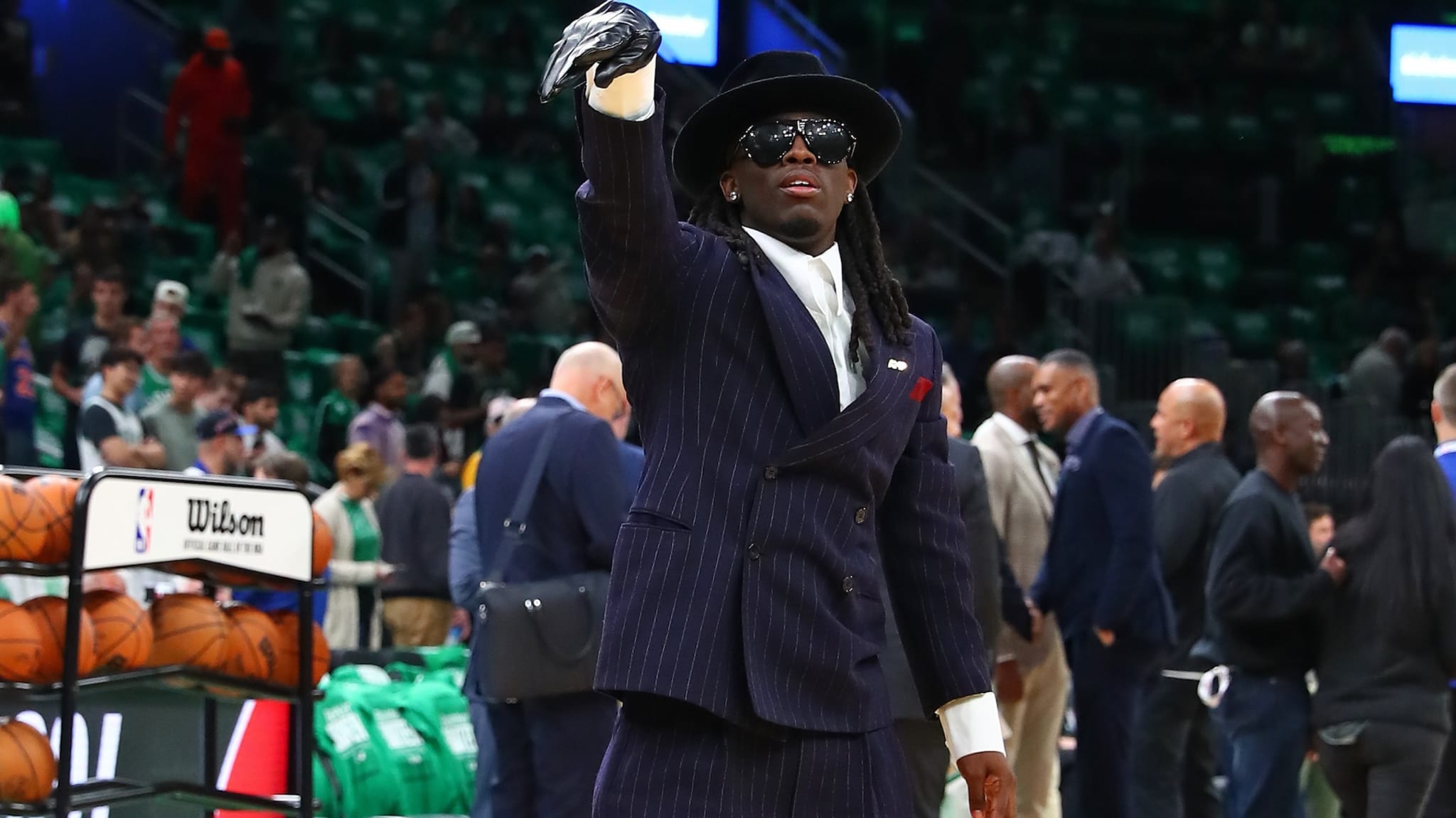Kai Cenat wearing pinstripe suit, top hat, large sunglasses, and leather glove on one hand at basketball game.