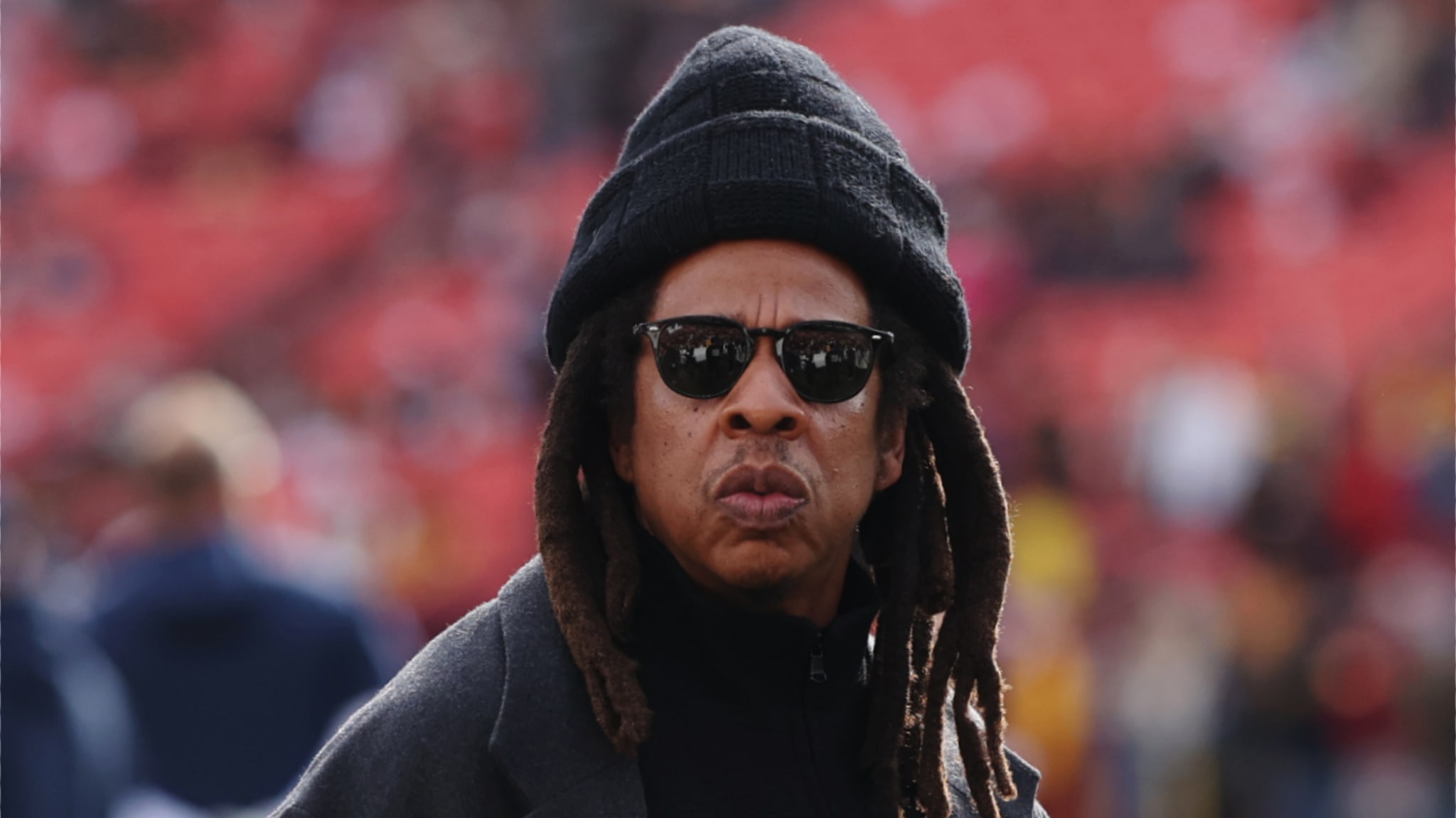 Jay-Z wearing sunglasses, a black beanie, and a dark coat stands outdoors with a blurred crowd in the background.