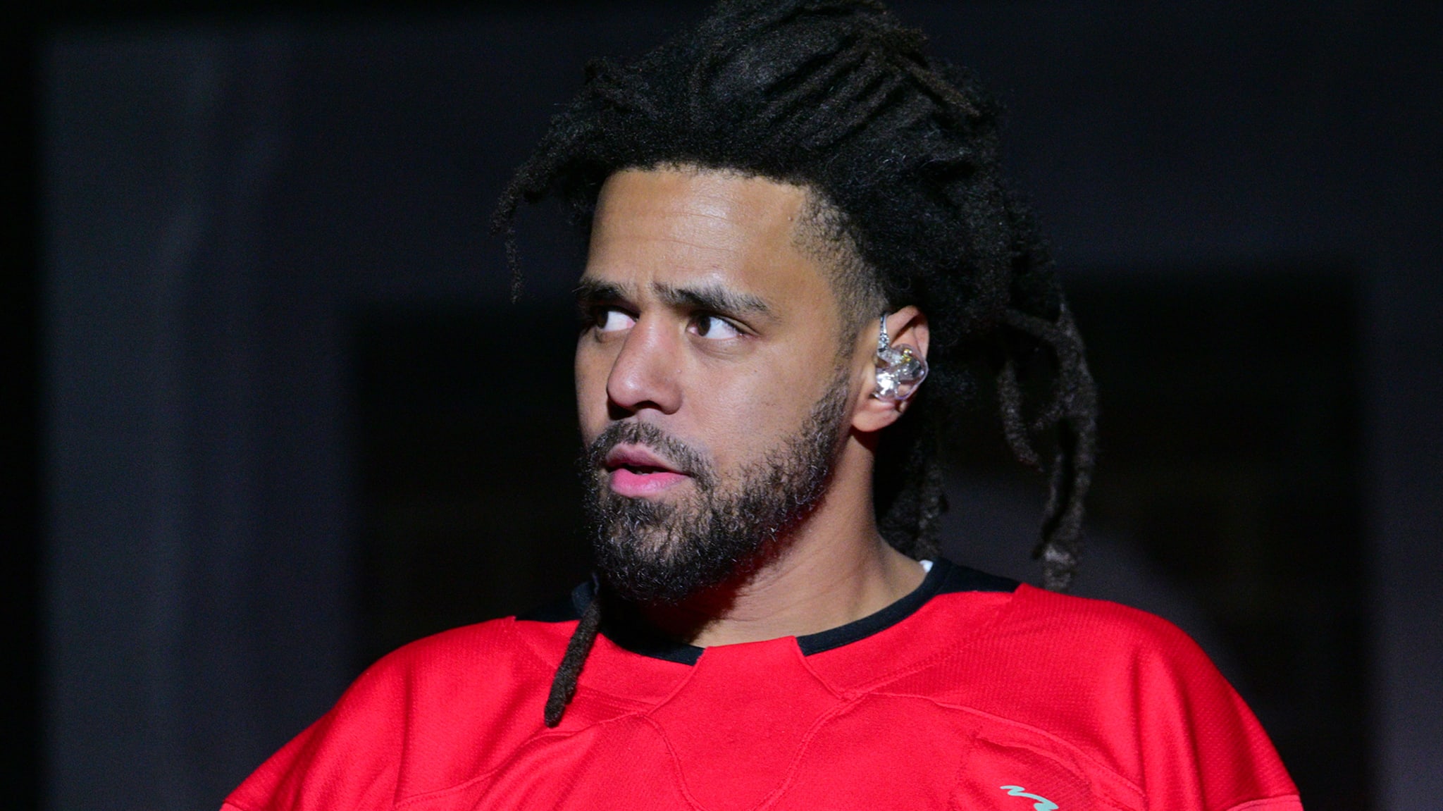 J. Cole performing on stage, wearing a red shirt, with an intense expression and dreadlocks.
