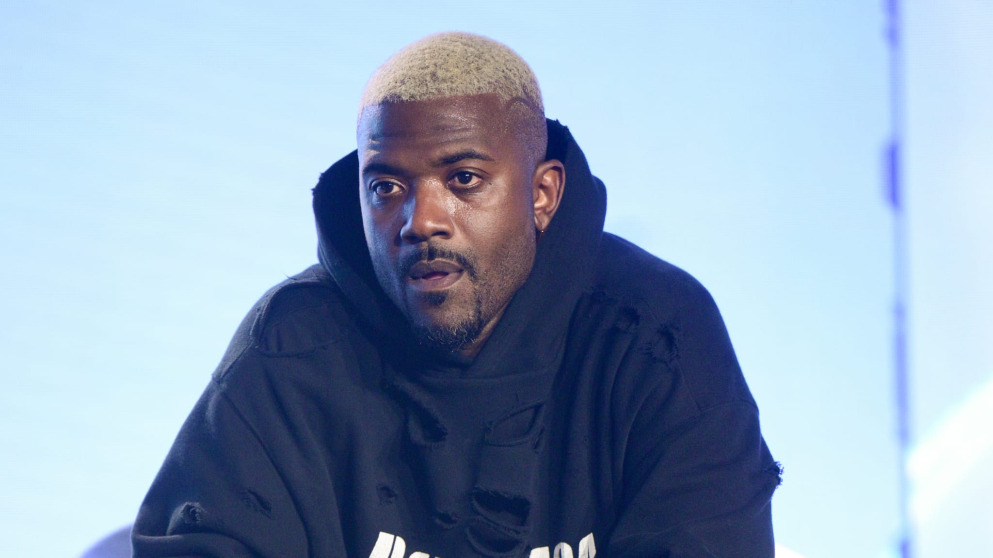A man with short, blonde hair wearing a black hoodie, looking attentively. The background is softly lit in blue tones.