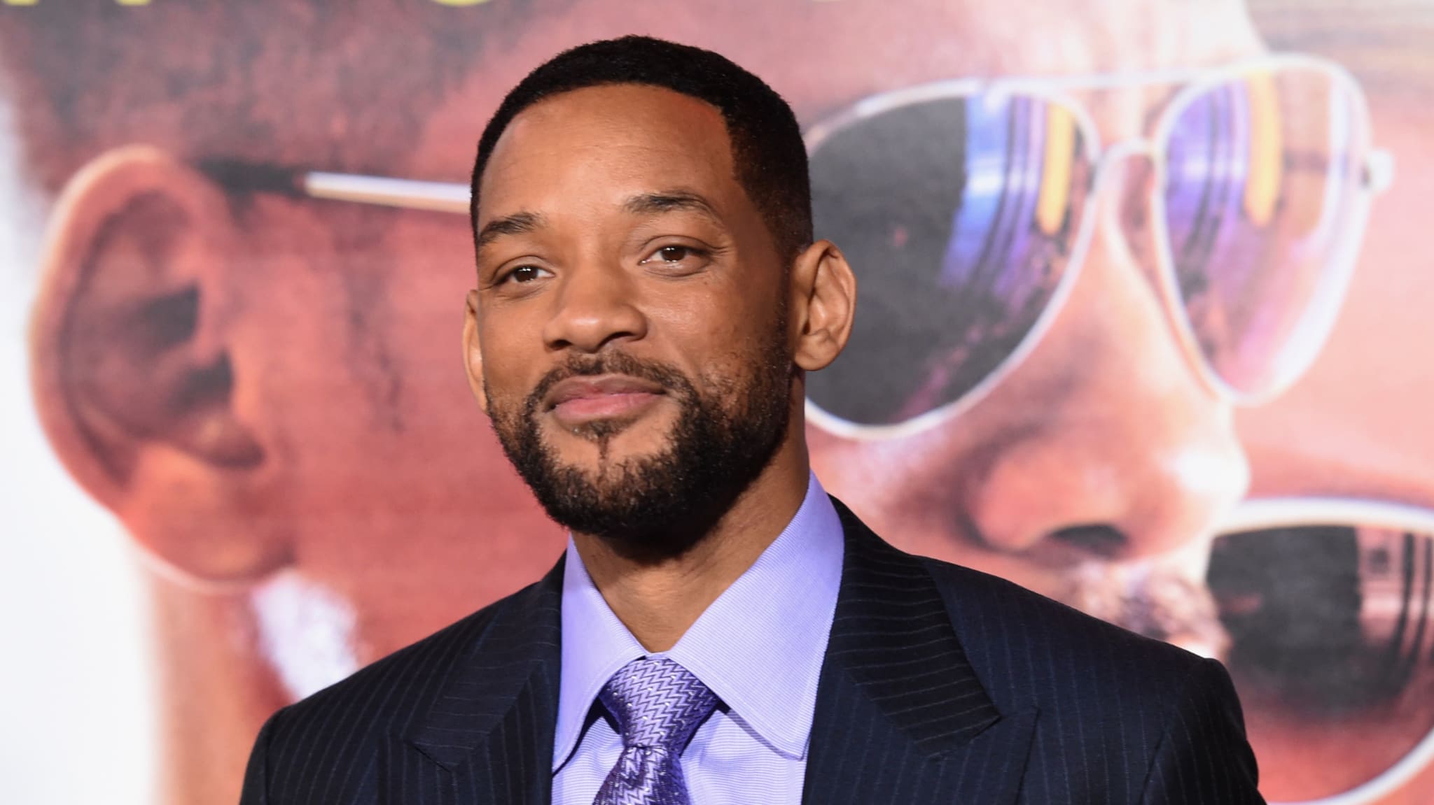 Will Smith in a suit and tie, smiling at an event, with a poster of him in sunglasses in the background.