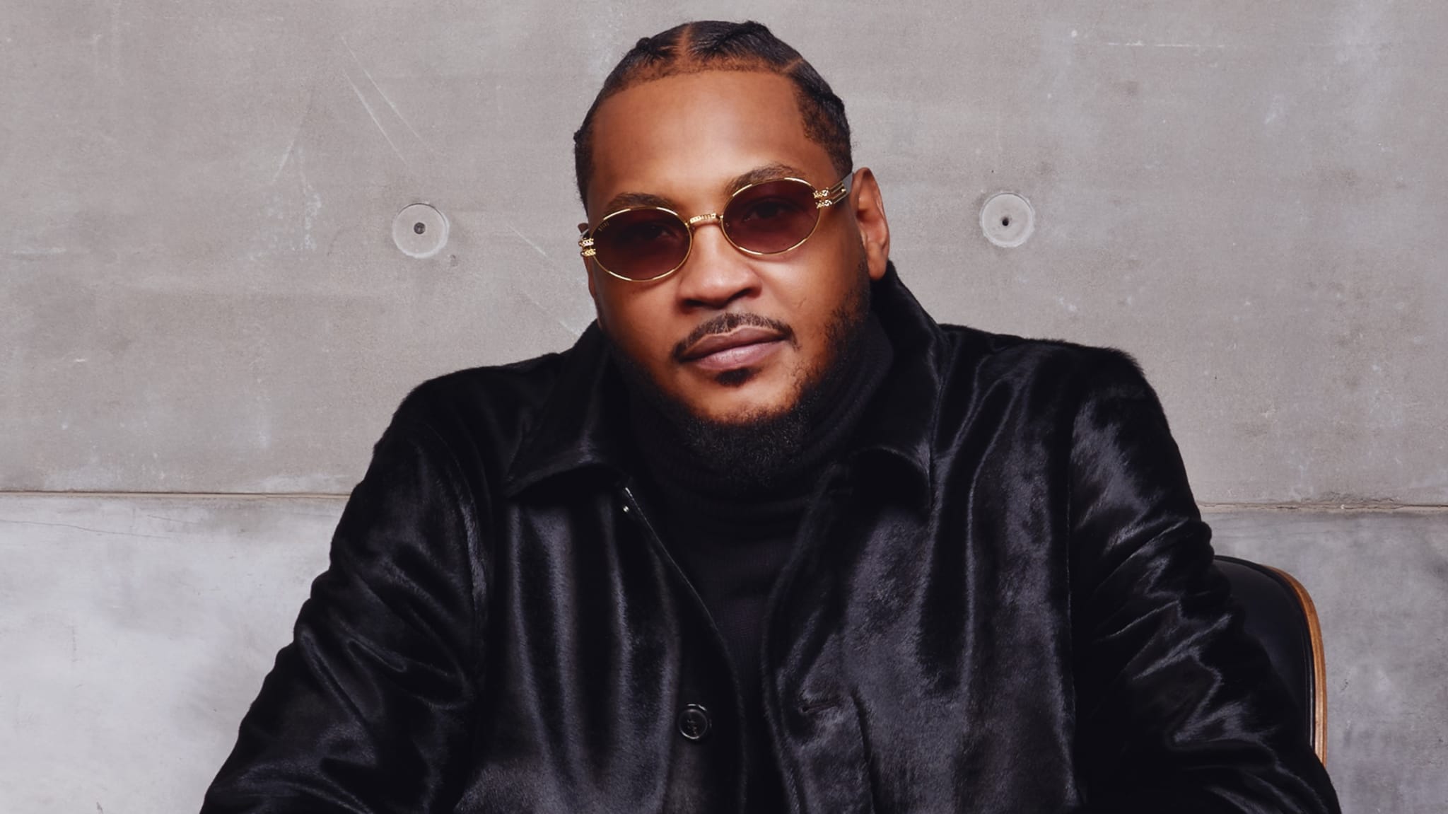 Carmelo Anthony wearing sunglasses and a black jacket, sitting against a concrete wall.