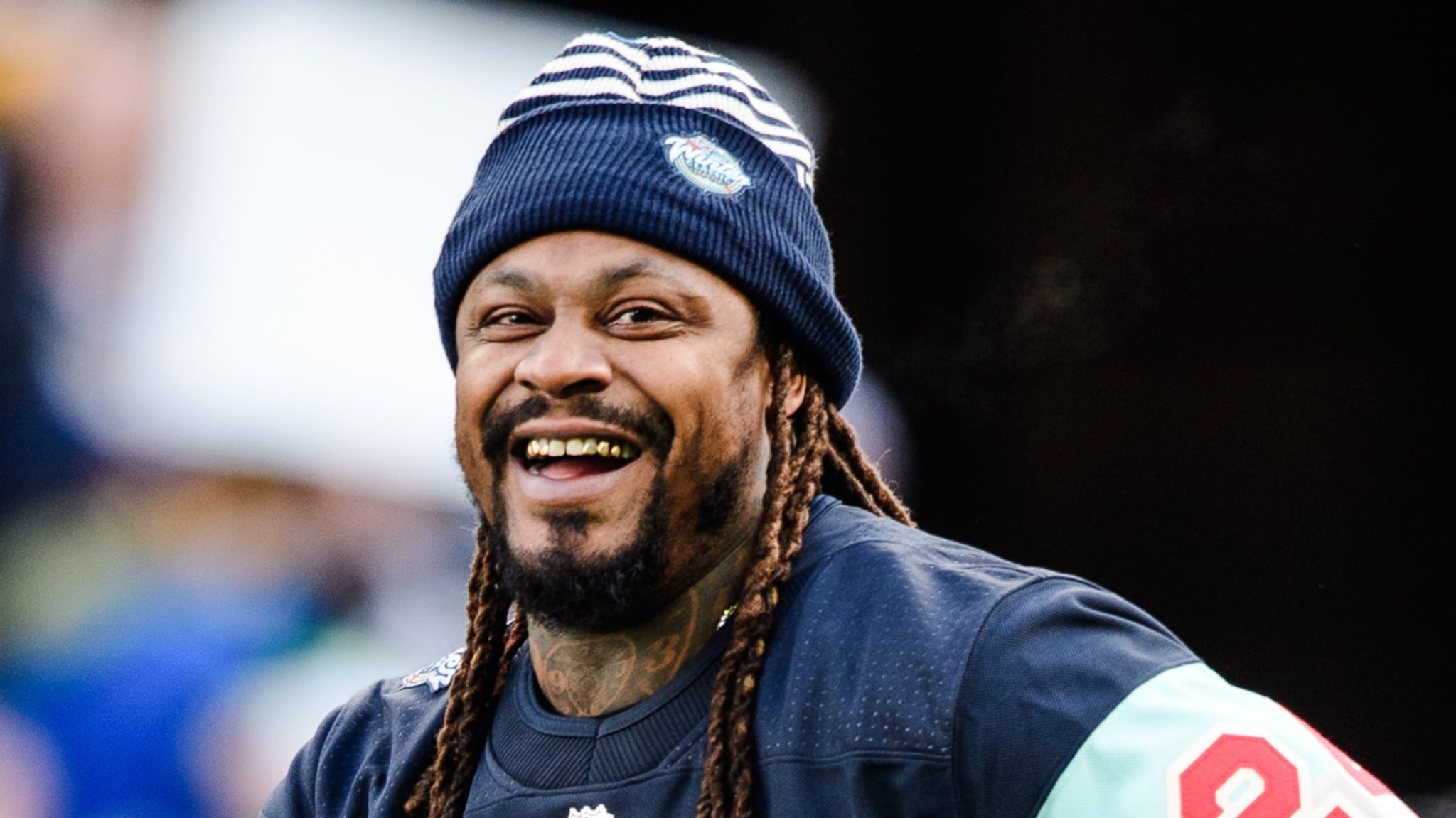 Marshawn Lynch, wearing a striped beanie, smiles broadly. He's dressed in a sports jersey, with long braids and a beard.