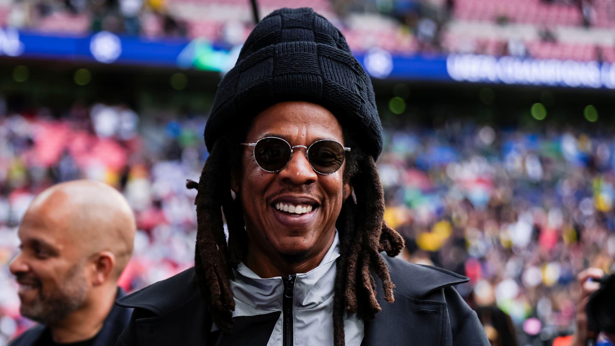 Jay-Z smiling at an event, wearing sunglasses and a black hat, with a crowd in the background.