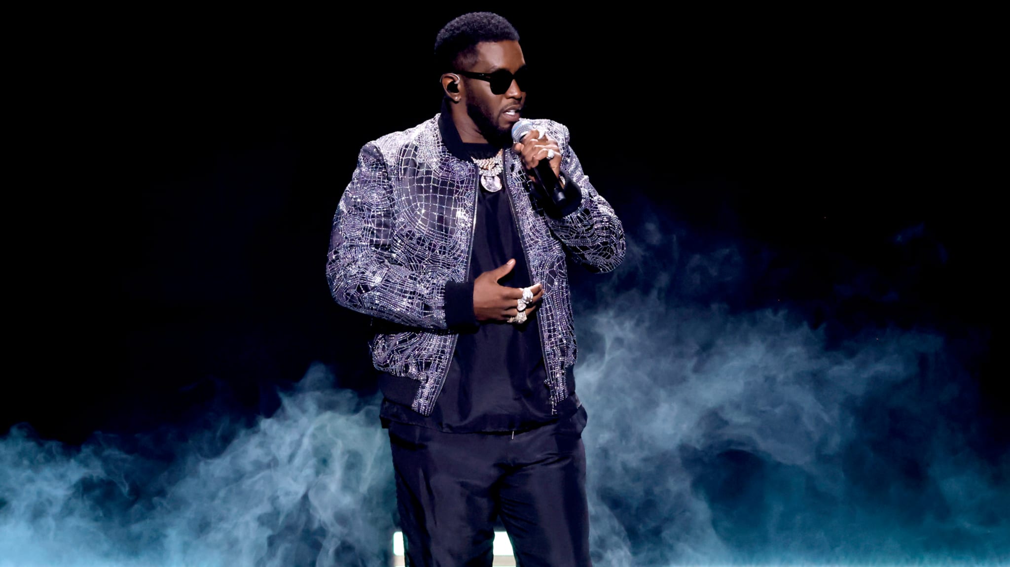 Sean "Diddy" Combs performing on stage, wearing sunglasses and a patterned jacket, surrounded by stage smoke.
