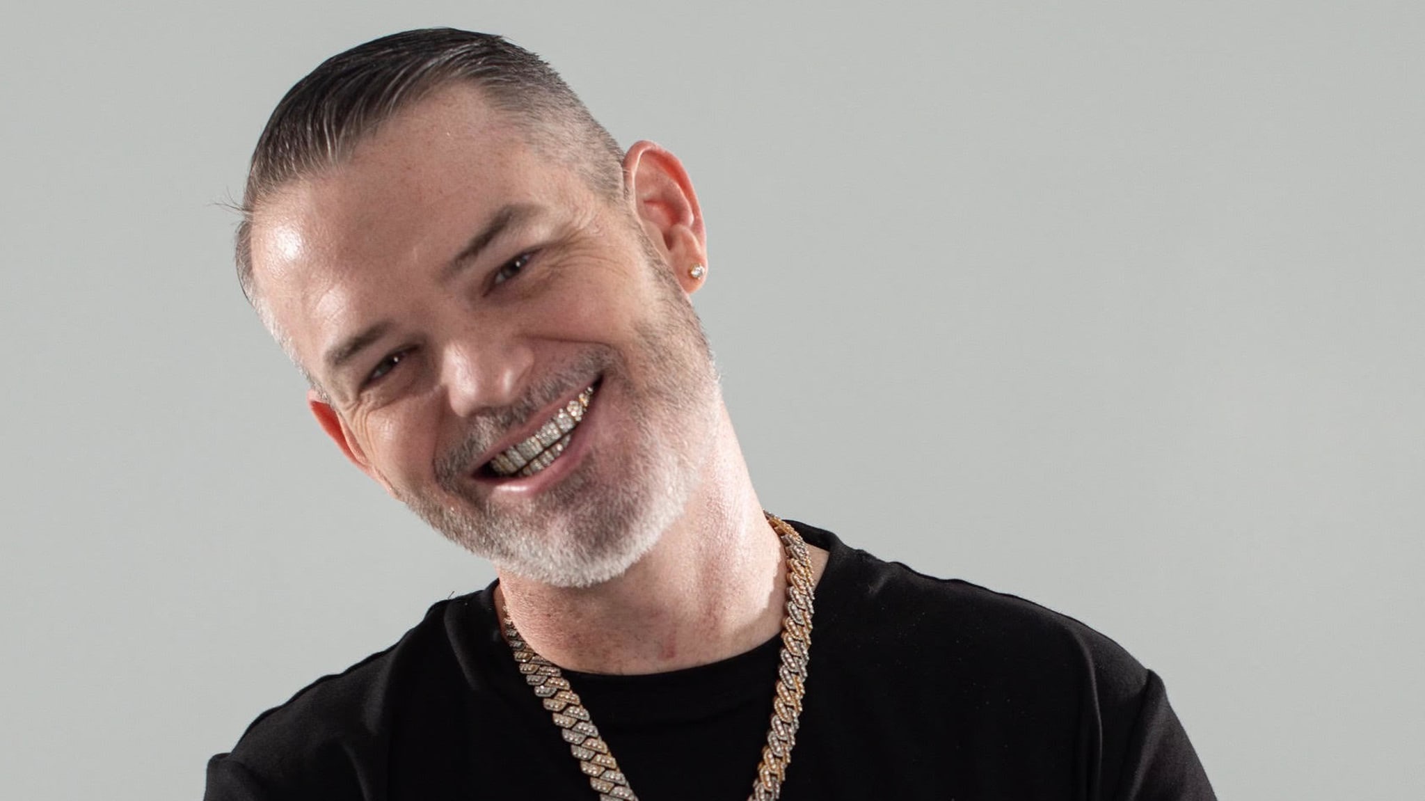 A man with a beard smiles, wearing a black shirt and gold chain. He has short, slicked-back hair and visible grills on his teeth.