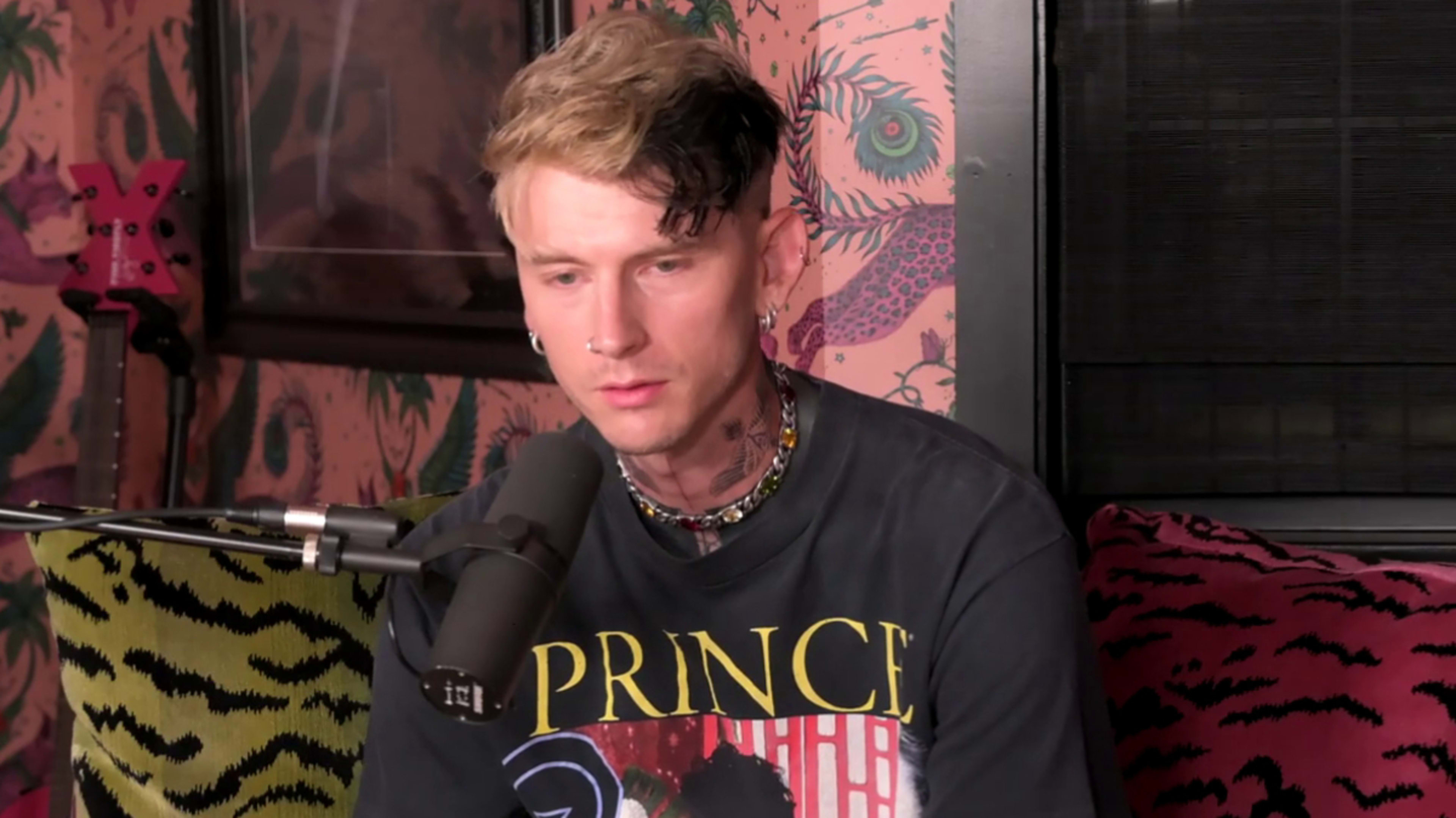 Machine Gun Kelly sits in front of a microphone wearing a Prince t-shirt and a beaded necklace. The background features patterned wallpaper