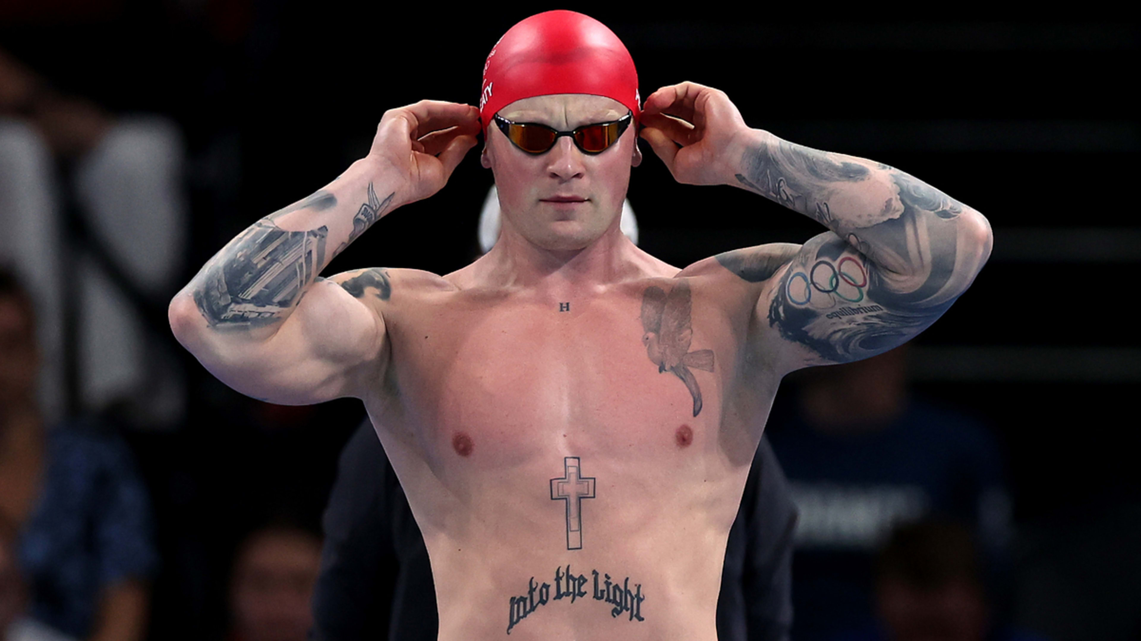 A male swimmer with tattoos adjusts his goggles. He wears a red swim cap and sunglasses, showcasing strong athletic build and focused expression before a race