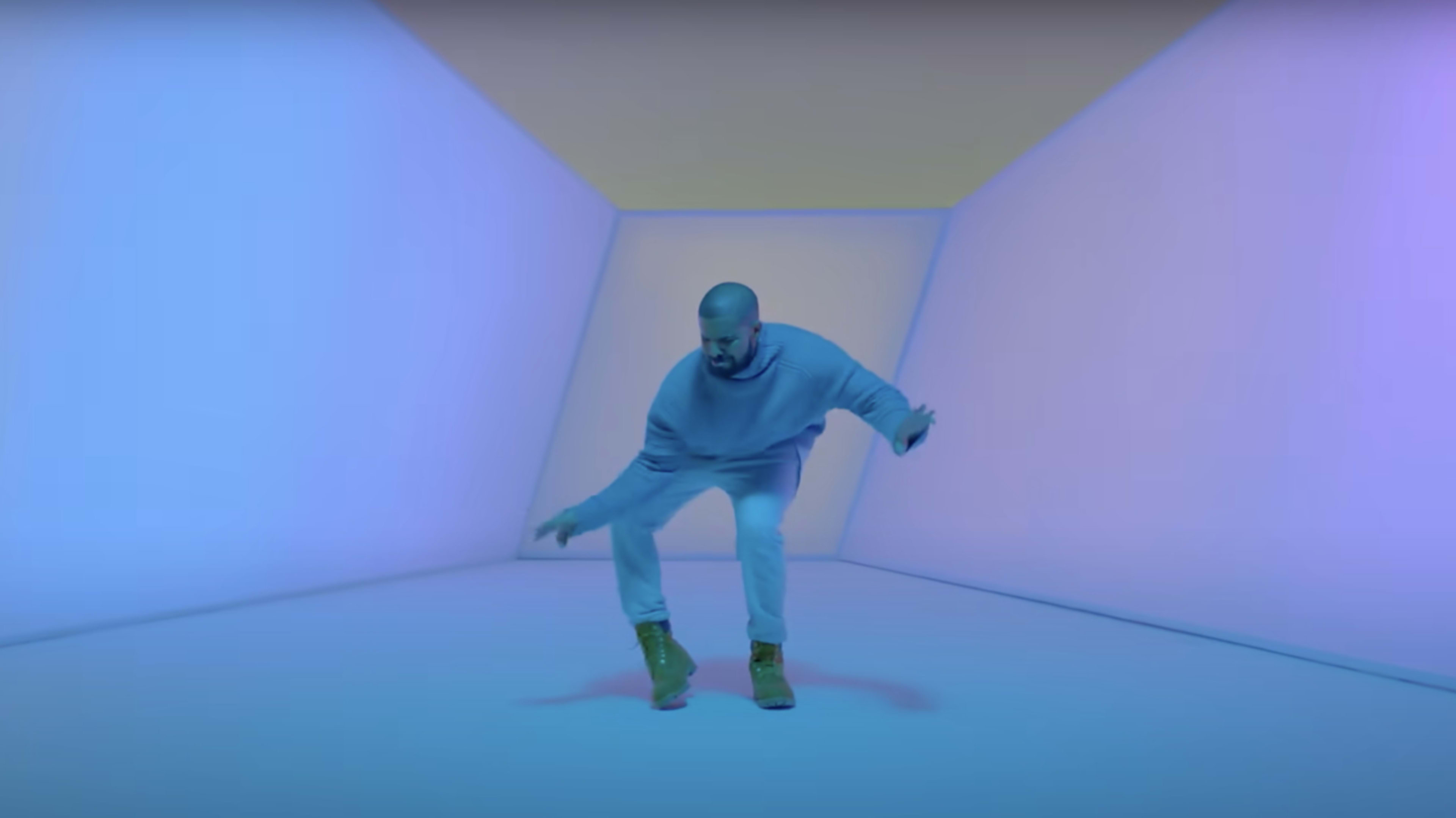 Drake dances in a futuristic, minimalistic room with soft lighting. He wears a casual outfit including a sweatshirt, jeans, and boots