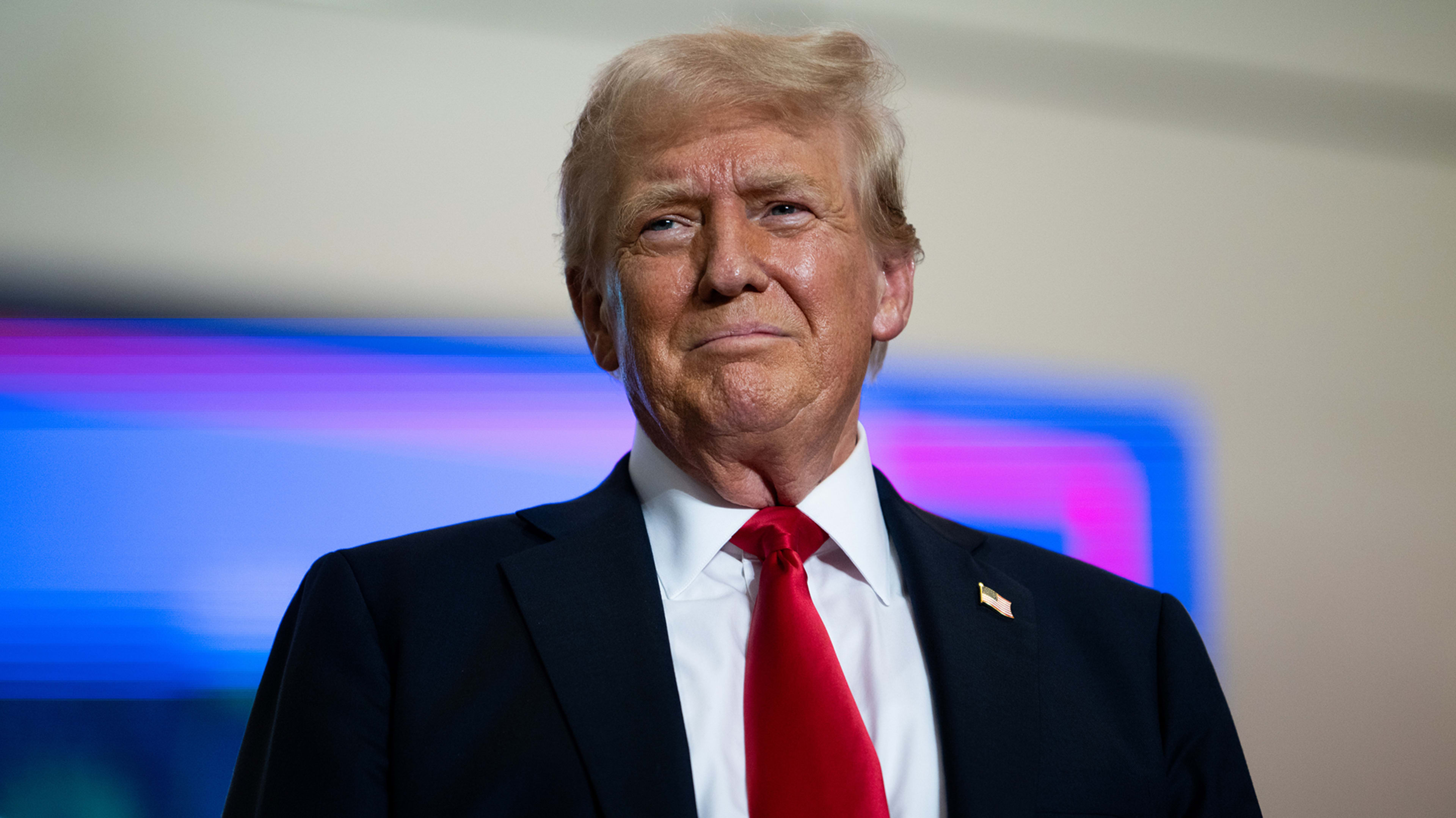 Donald Trump wearing a suit with a white dress shirt and a red tie, standing in front of a blurred background