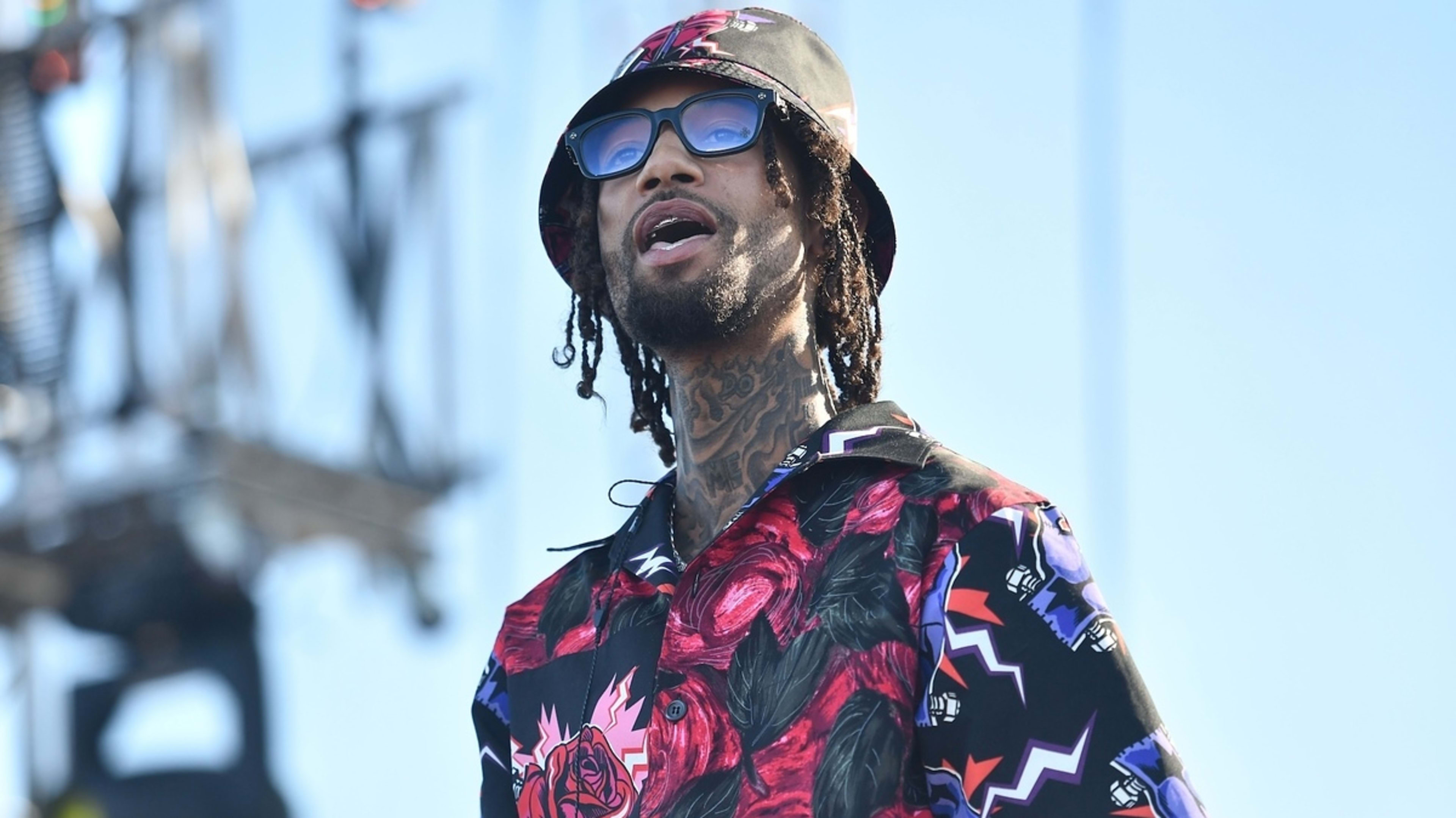 PnB Rock wearing a patterned bucket hat, dark sunglasses, and a black shirt adorned with colorful designs