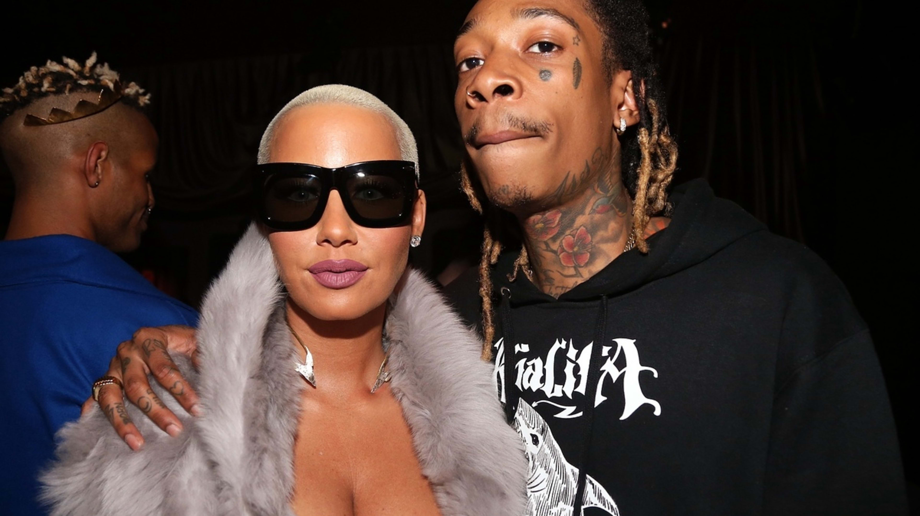 Amber Rose wearing large sunglasses and a fur-trimmed coat, stands beside Wiz Khalifa, who wears a hoodie with tattoos visible on his face