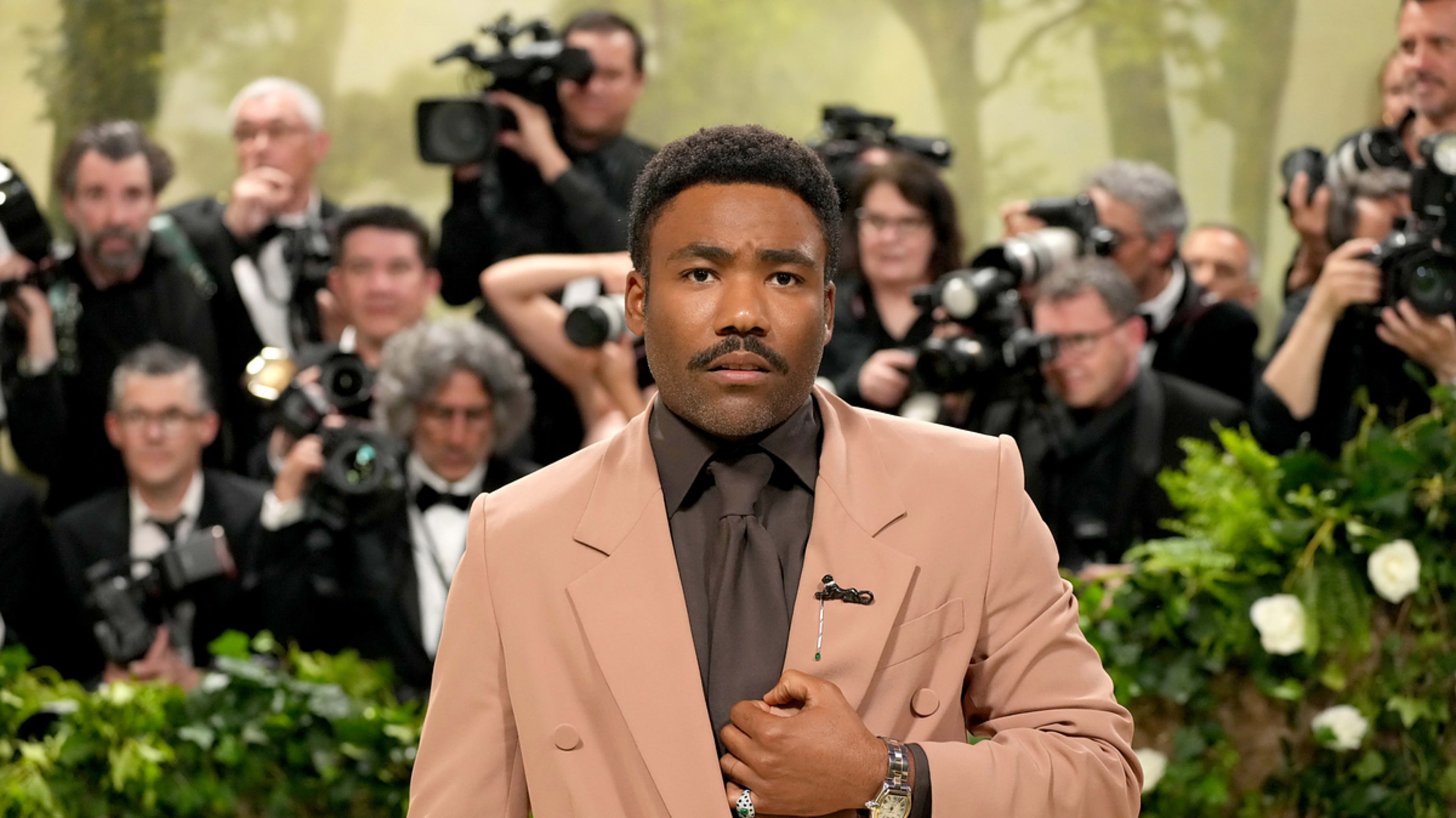 Photographers capture Donald Glover on the red carpet at an event. He wears a tan suit with a dark shirt and tie