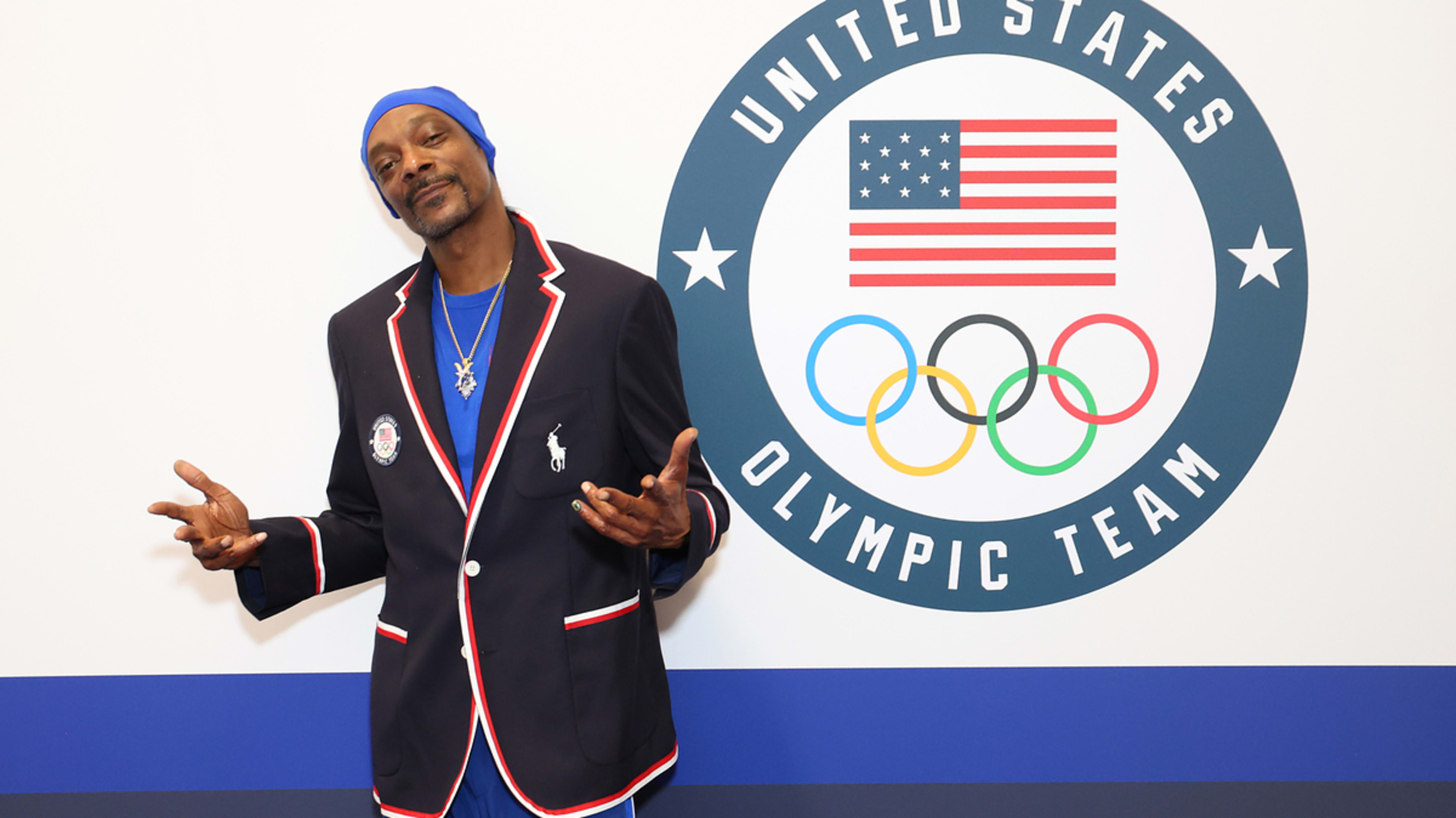 Snoop Dogg in athletic pants, a shirt, and a blazer with Olympic Rings and a U.S. emblem, stands in front of a backdrop with the United States Olympic Team logo