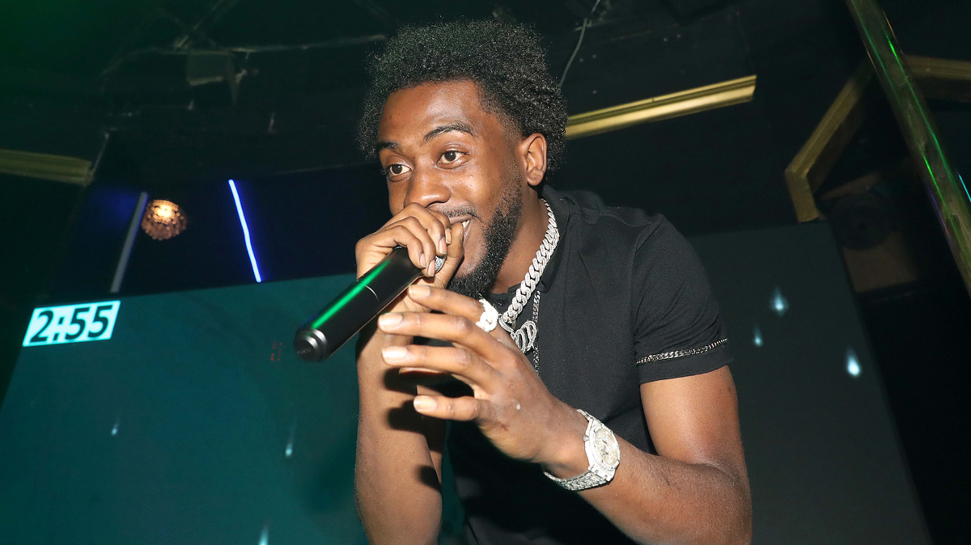 Desiigner performing on stage, holding a microphone with an energetic expression. He is wearing a simple black t-shirt, a silver chain necklace, and a matching watch