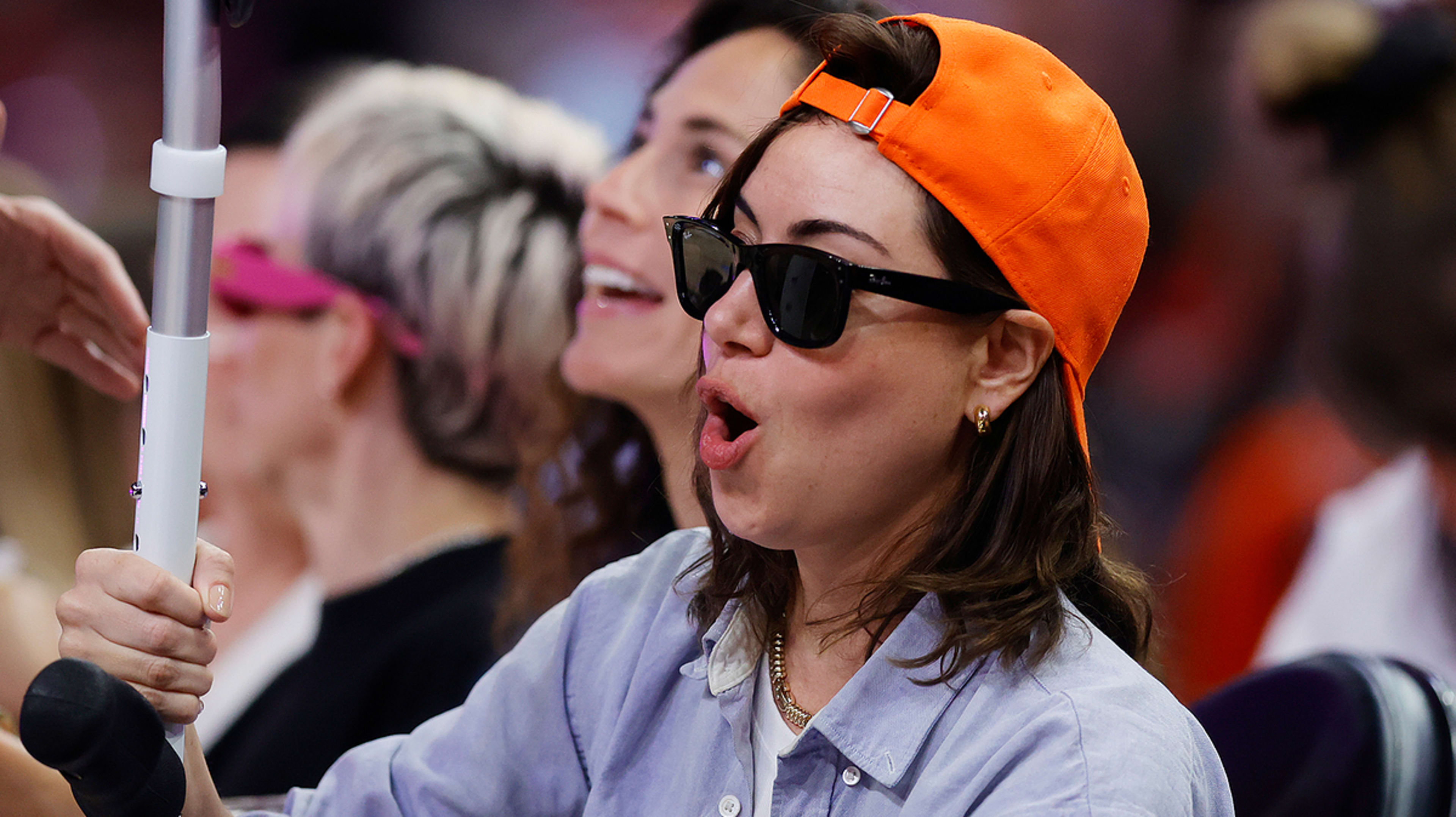 Aubrey Plaza wears sunglasses and an orange cap, holding a walking cane and reacting excitedly at an event. Other individuals are seated nearby