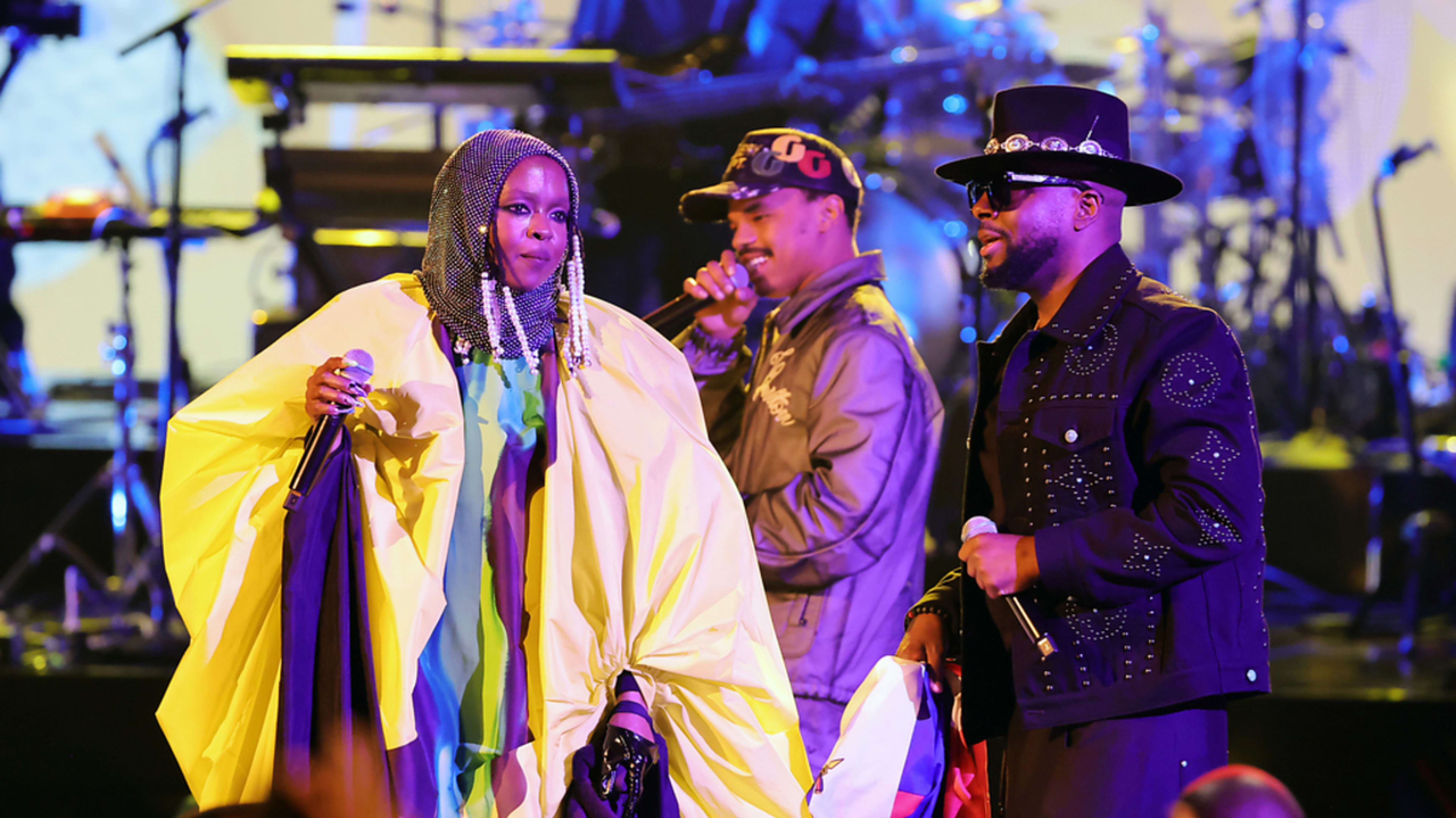 Lauryn Hill, Pras Michel, and Wyclef Jean perform on stage together. Lauryn wears a flowing, draped outfit with a beaded headdress; Pras and Wyclef wear streetwear