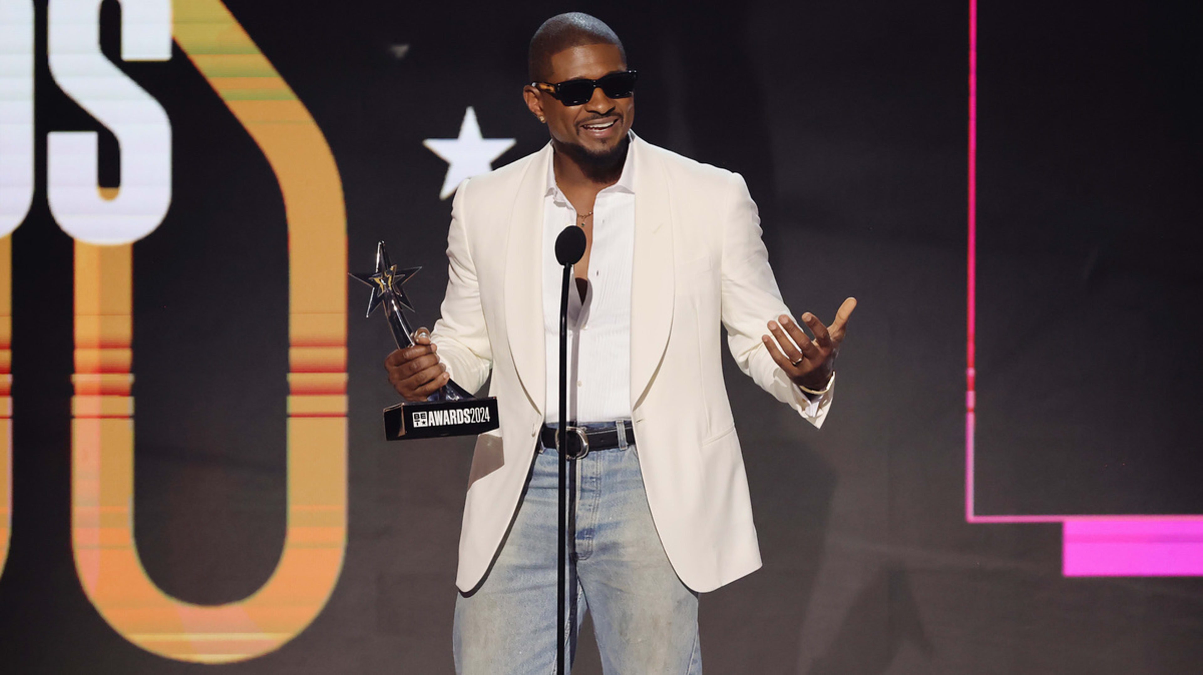 Usher in a white jacket and jeans, holding an award, speaks on stage at an event