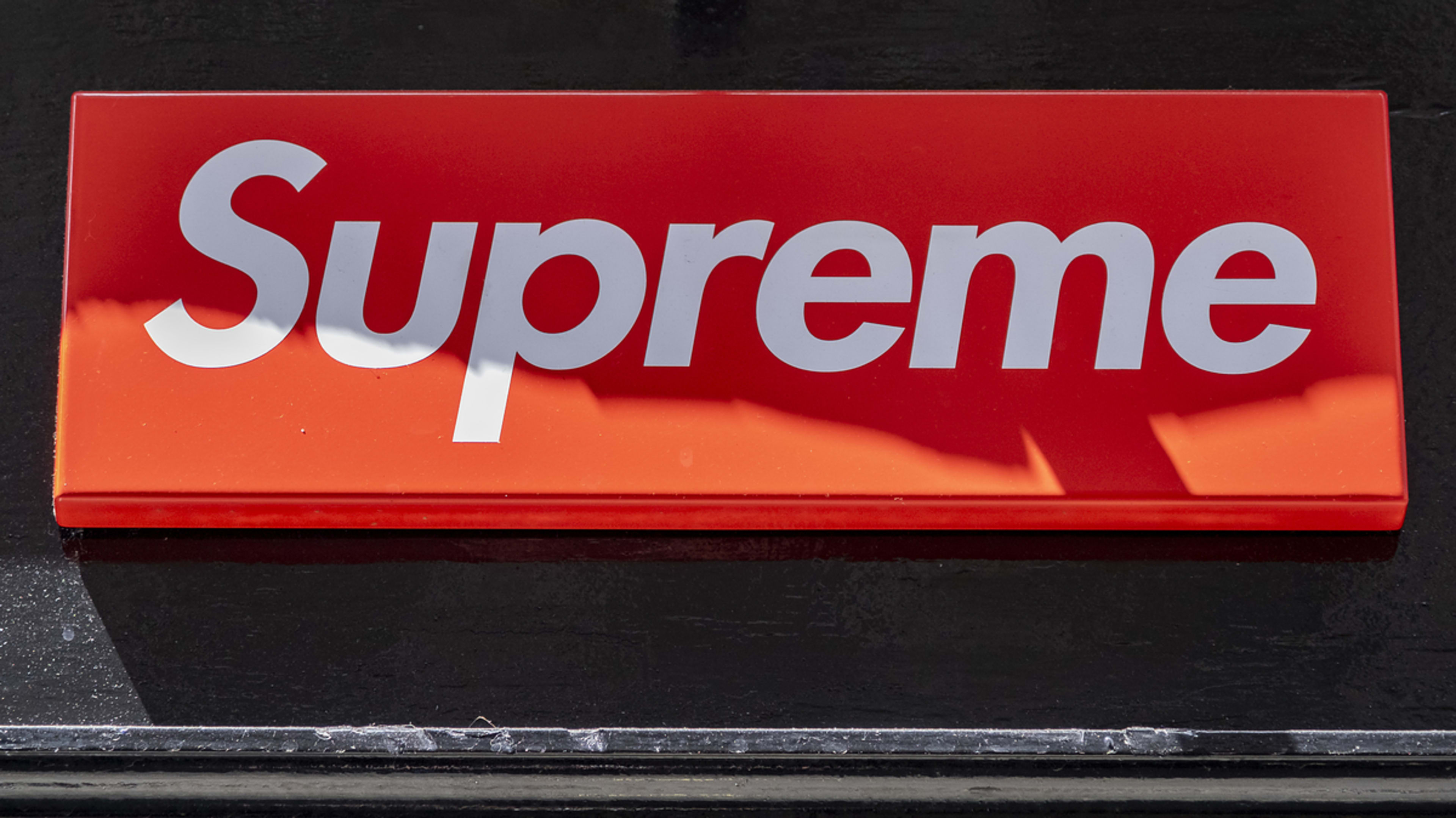 A Supreme store sign with bold white text on a rectangular background. The sign features the brand name "Supreme" prominently