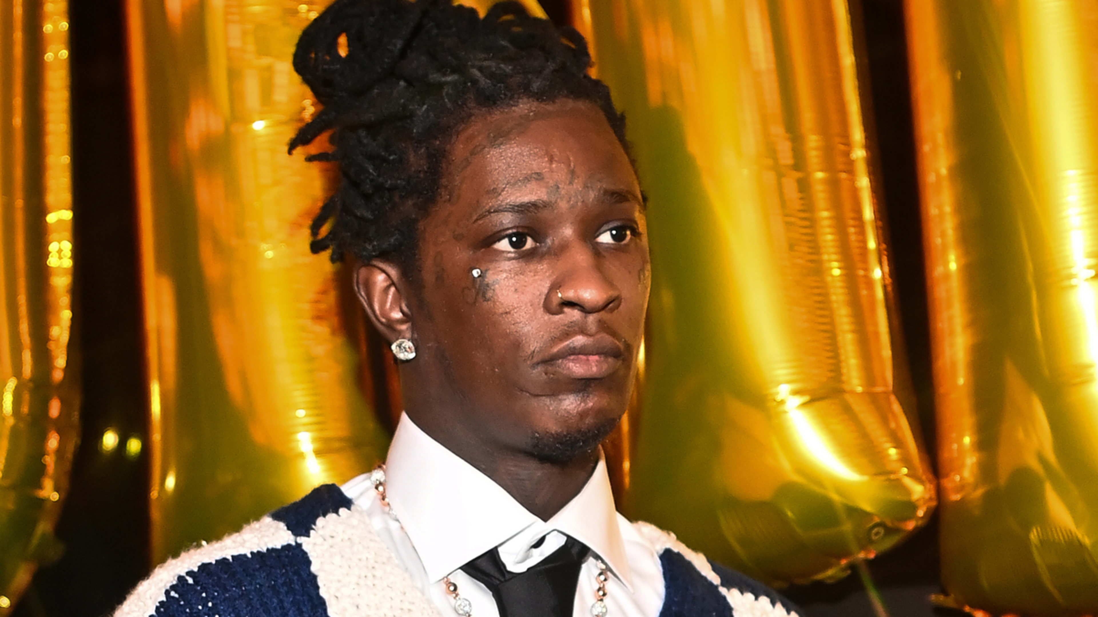 Young Thug wearing a white shirt, black tie, and knitted sweater, standing in front of gold balloons