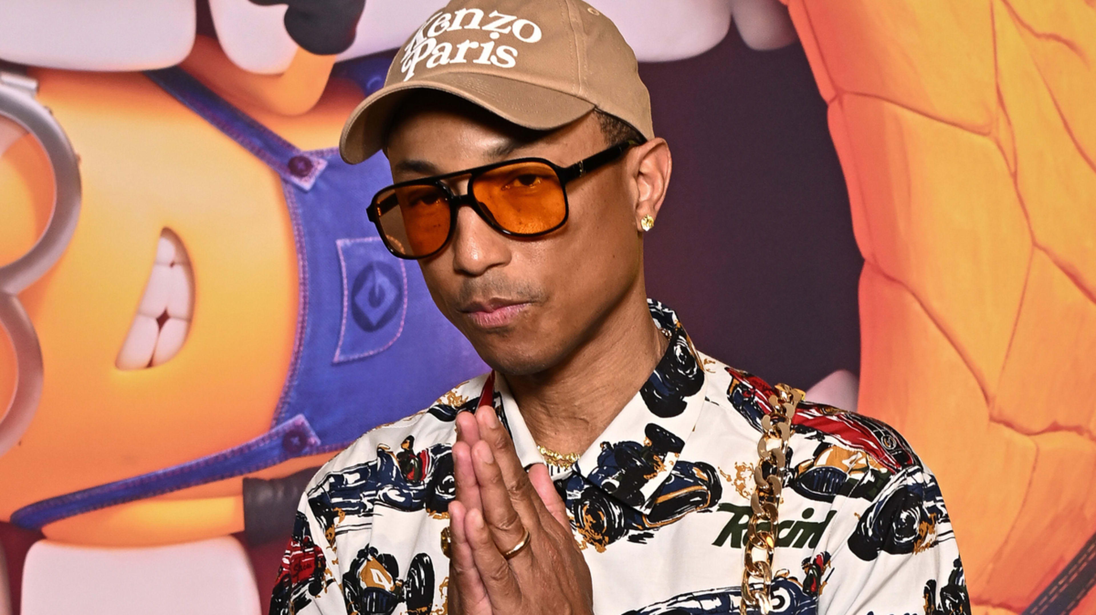 Pharrell Williams wearing a patterned shirt, orange-tinted glasses, and a cap with "Kenzo Paris" written on it, posing with his hands together in front of a colorful background