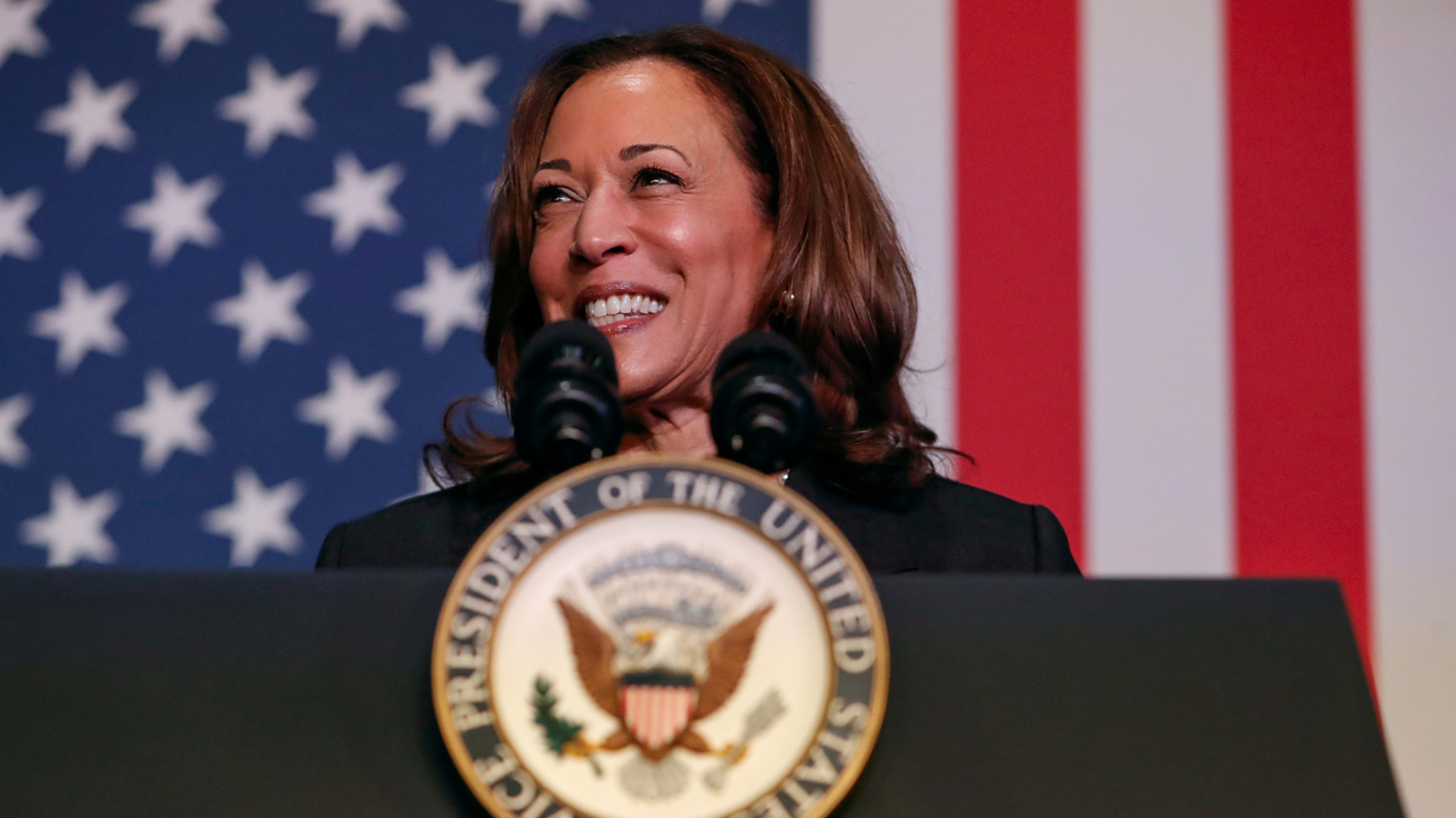 Kamala Harris smiling and speaking at a podium with the United States presidential seal, in front of an American flag backdrop