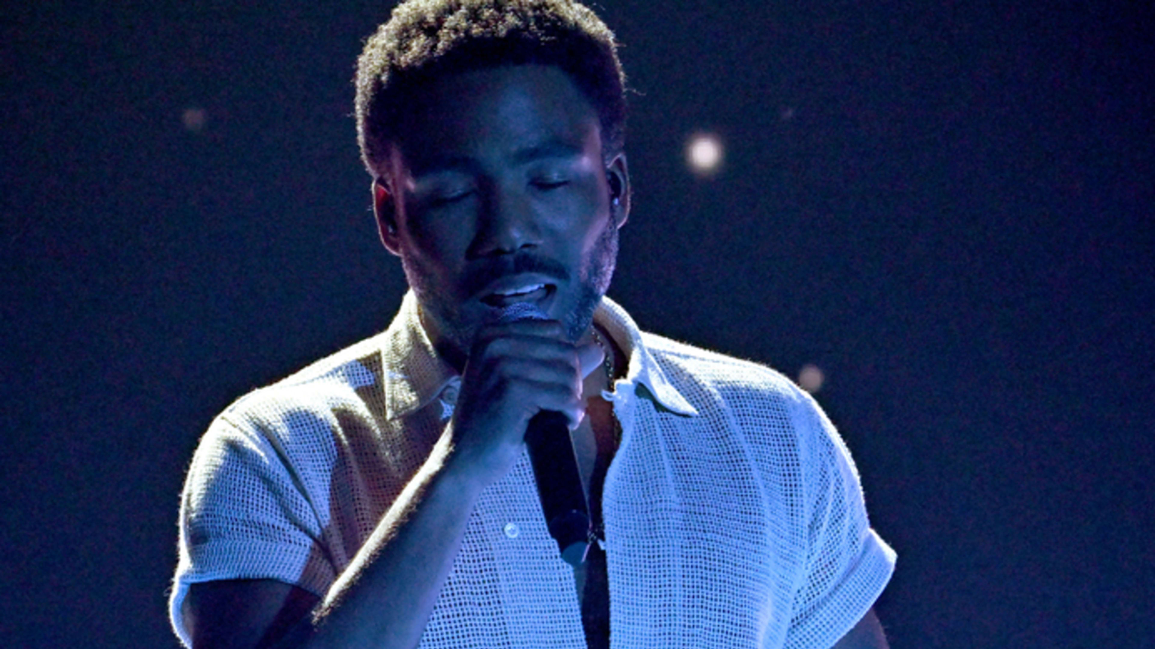 Donald Glover performs on stage, wearing a casual open-neck shirt and holding a microphone against a dark background with scattered lights