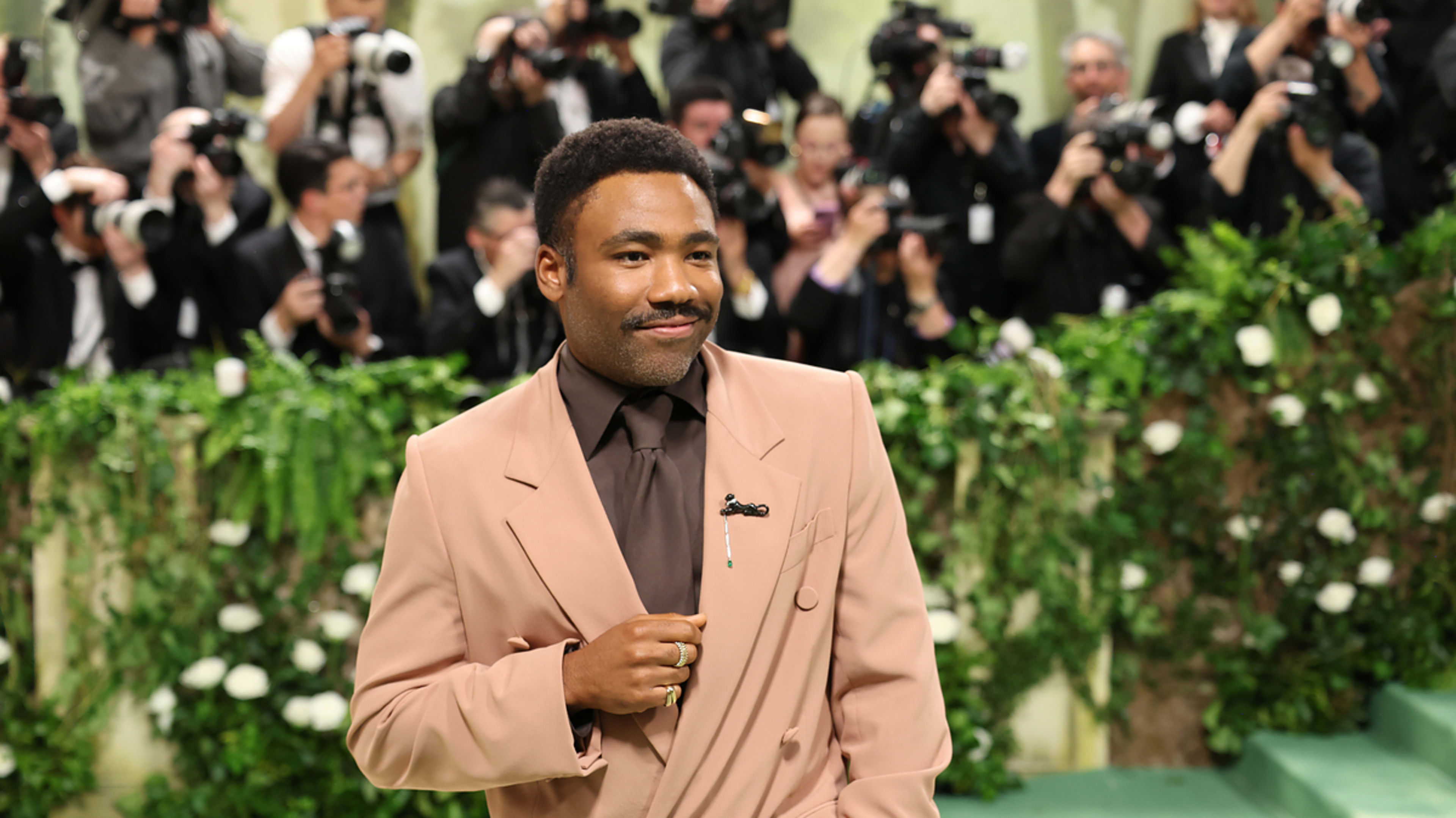 Donald Glover on a red carpet in a tan suit and dark shirt, smiling for photographers at a formal event