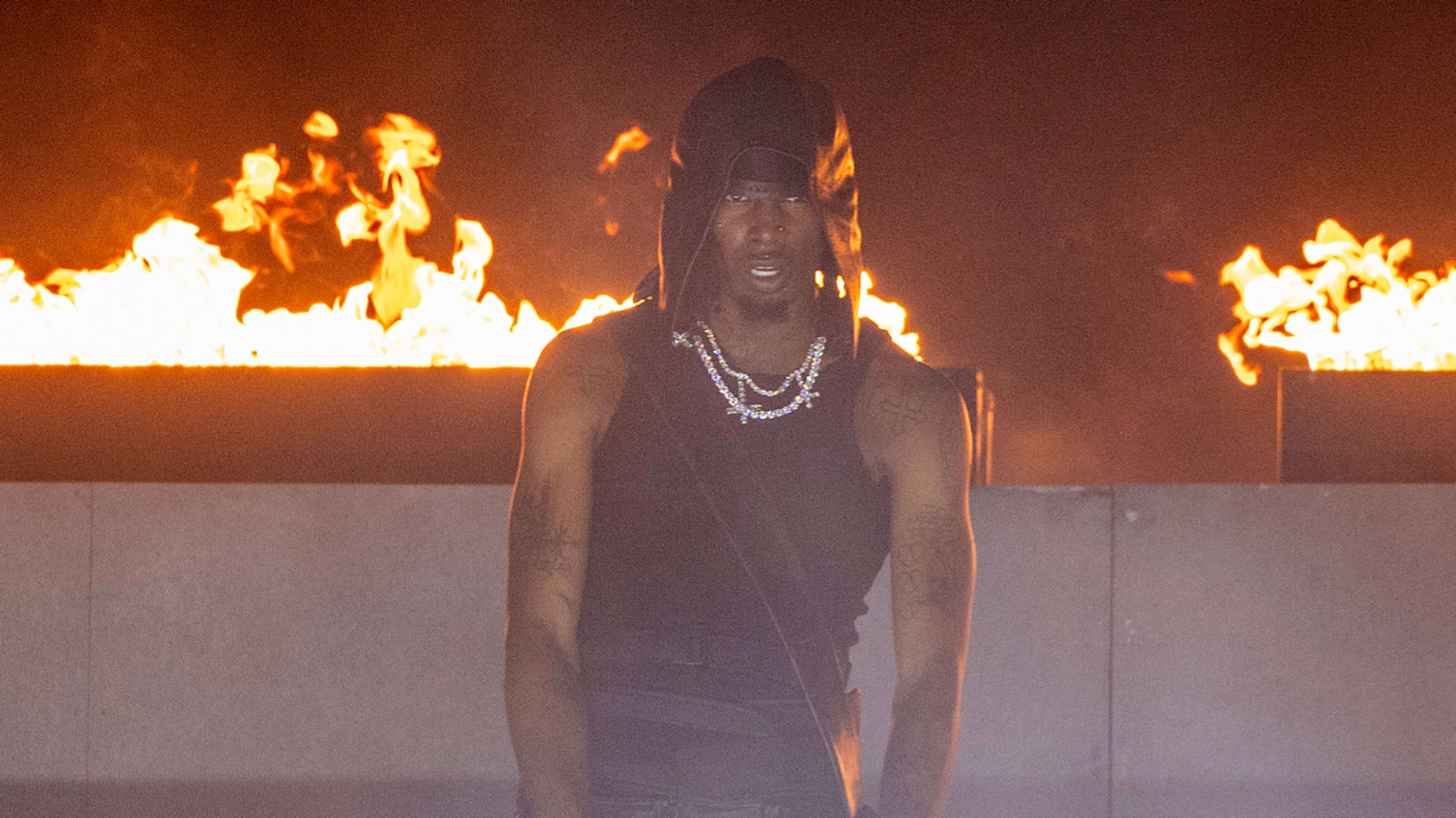 Playboi Carti performs on stage wearing a black tank top and black head covering, with fire blazing in the background