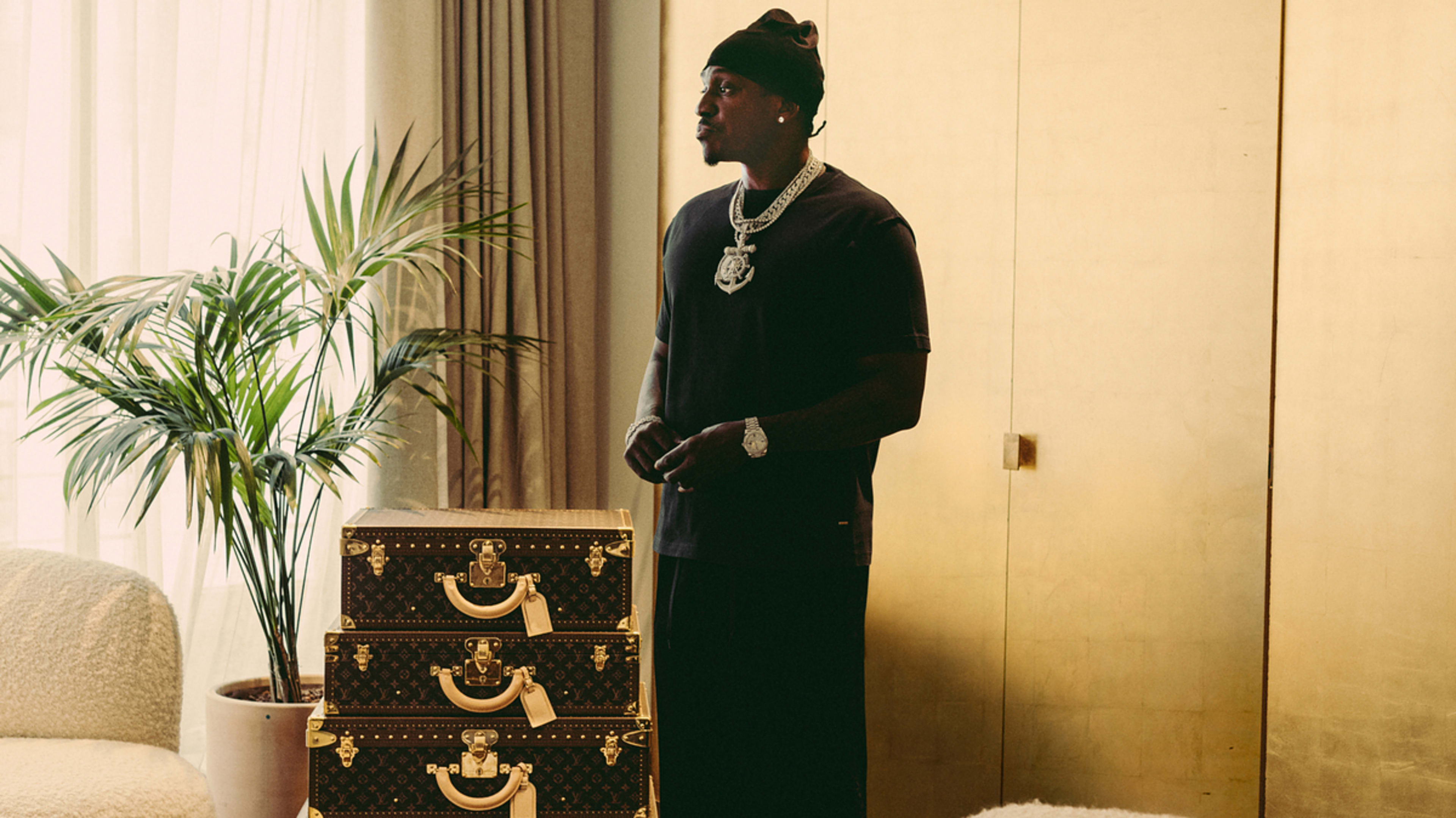 Person stands indoors near stacked designer luggage, wearing a black t-shirt, black hat, and large jewelry in a stylish, relaxed pose