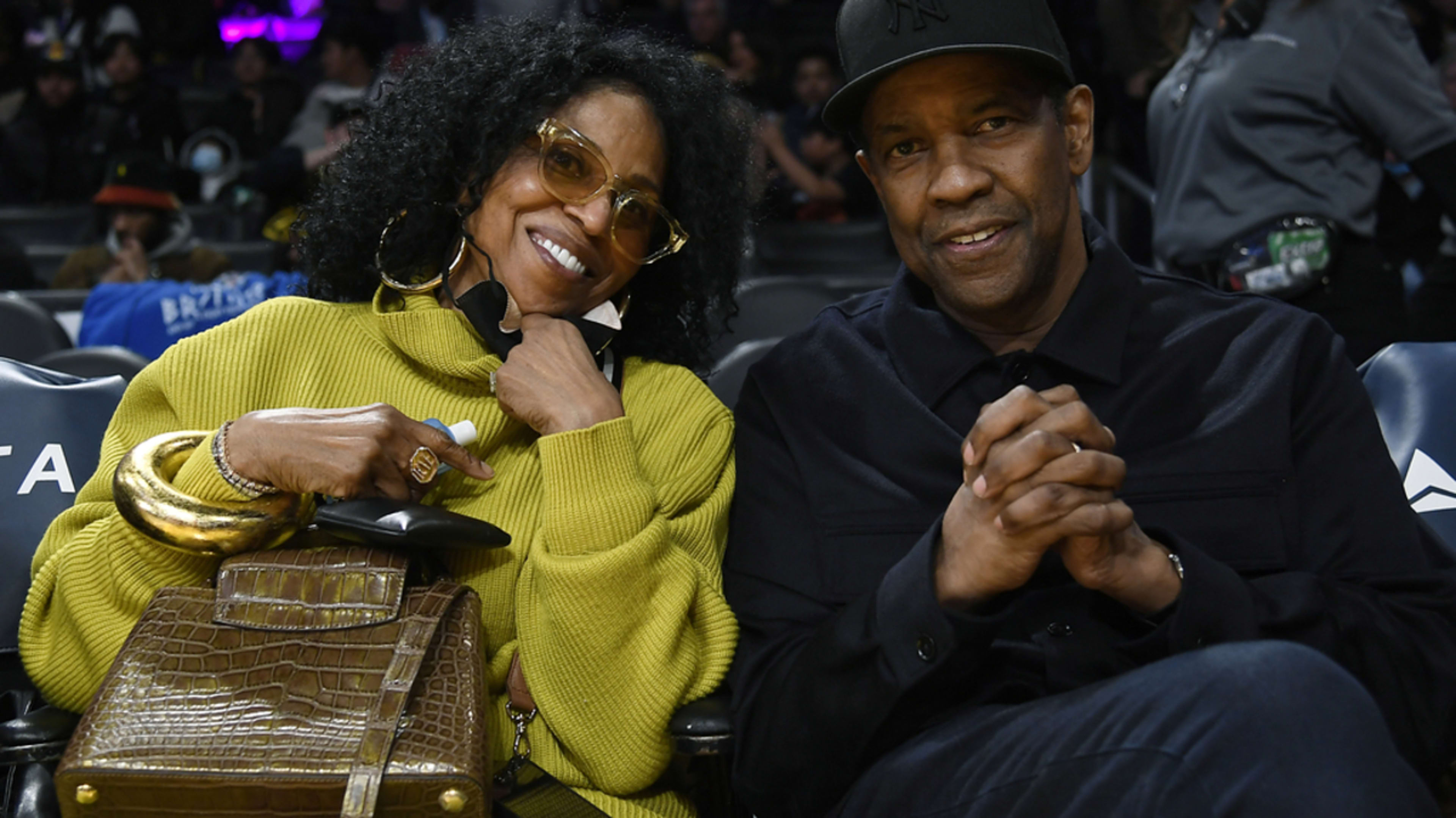 Diana Ross, in a cozy chic outfit, and Denzel Washington, in a casual black ensemble, sit side by side at a crowded event, smiling at the camera