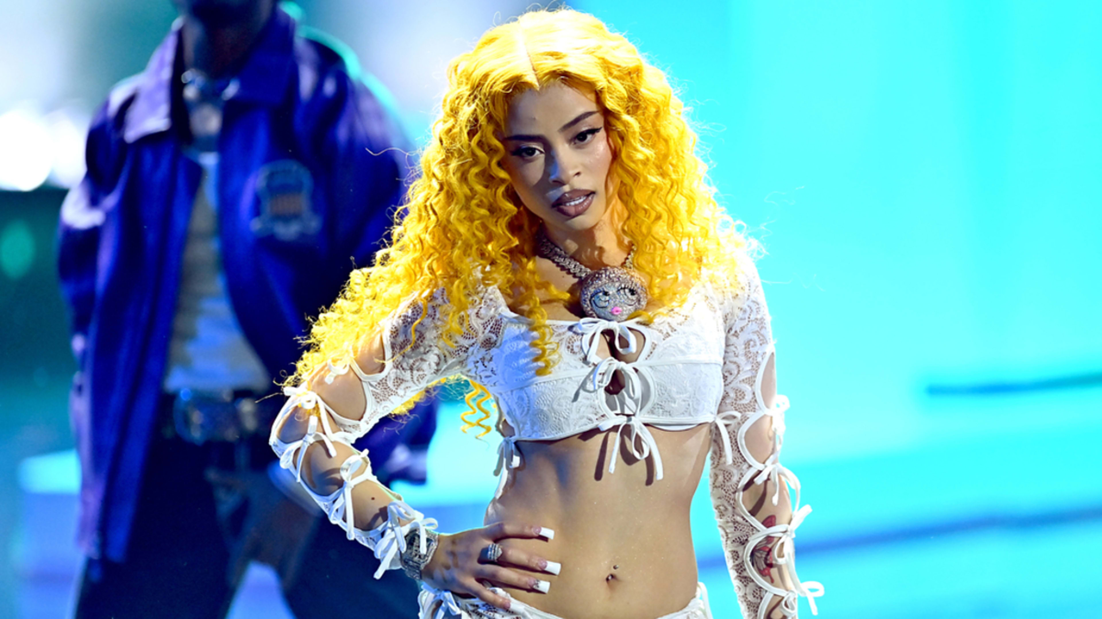 A person performs on stage wearing a lace crop top and shorts. A figure in the background is partially visible. Names are not provided