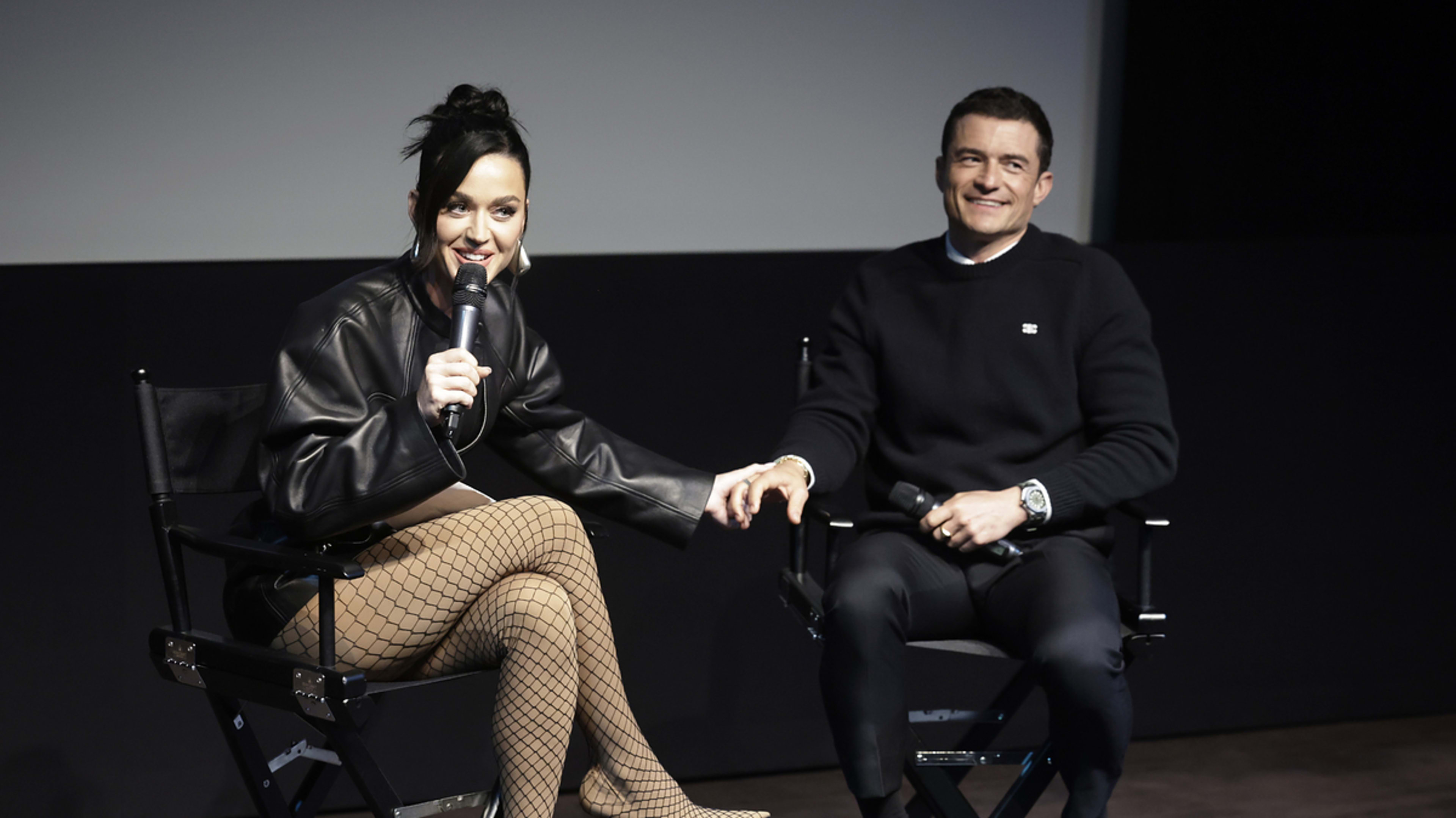 Katy Perry and Orlando Bloom sitting on a stage, holding hands. Katy wears a black leather jacket and fishnet stockings, while Orlando is in a black sweater