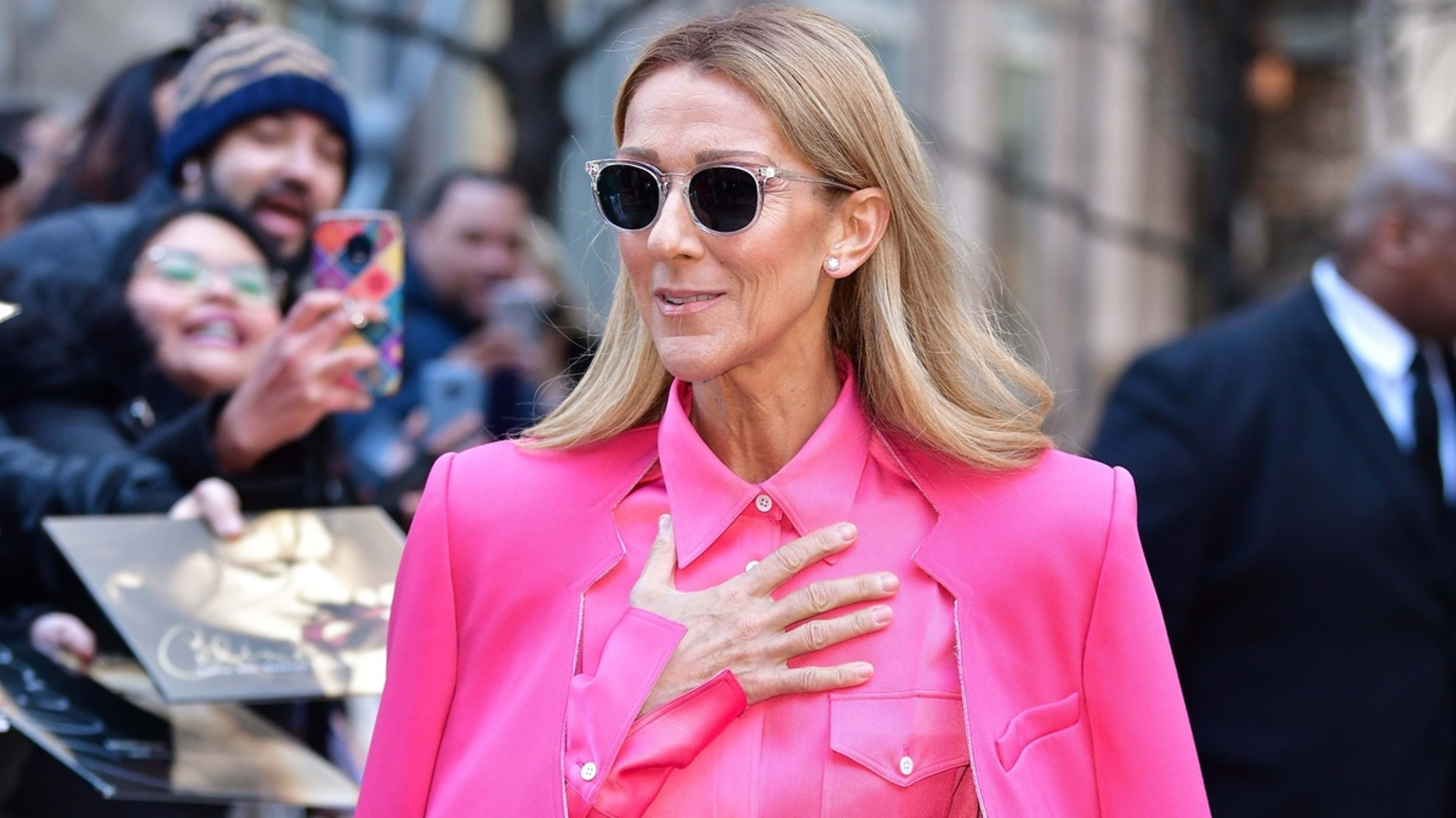 Celine Dion, dressed in stylish, bold clothing, walks outside while fans in the background take photos