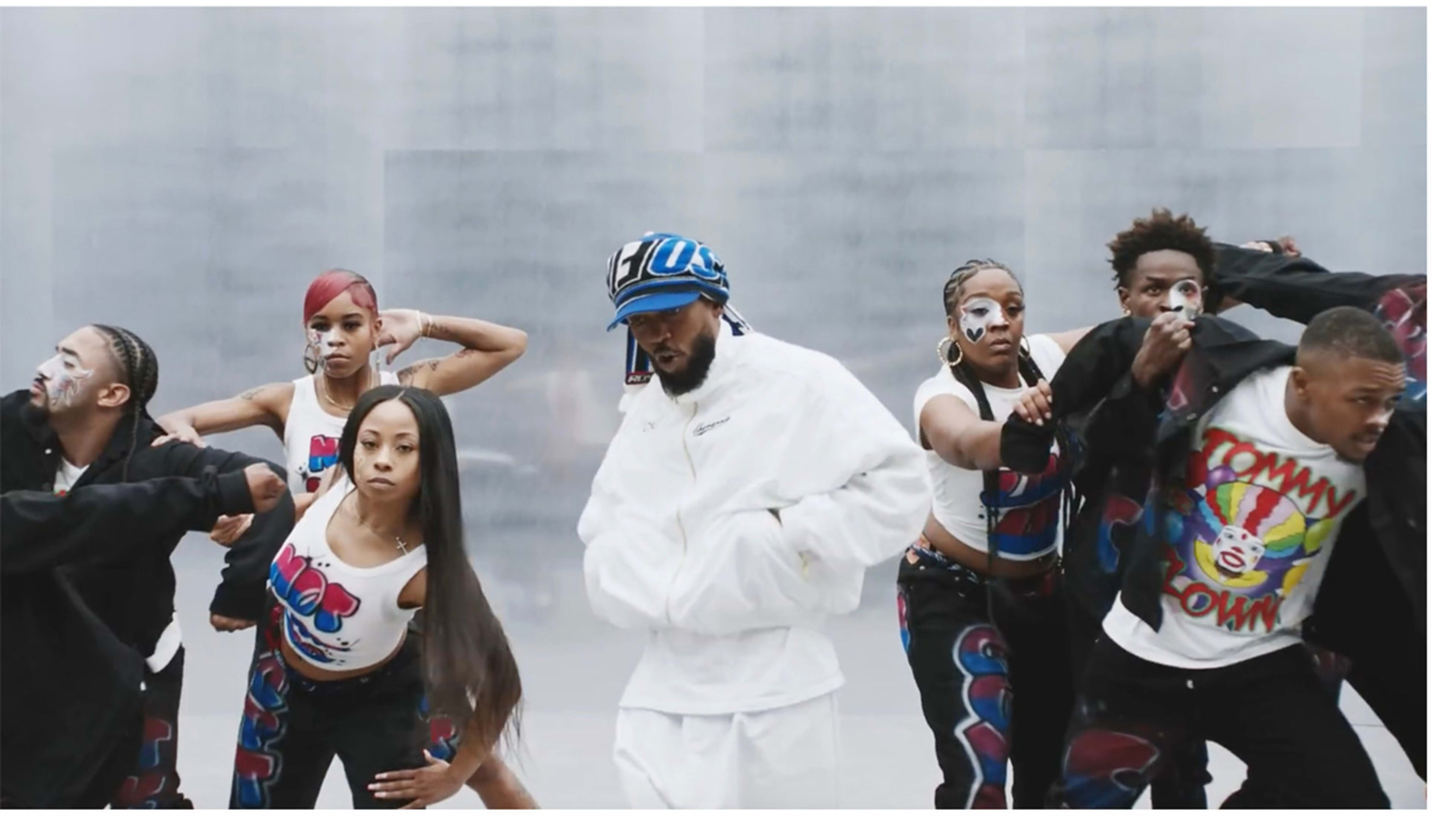 Kendrick Lamar in a white tracksuit and blue hat dances with a group of dancers in matching outfits, featuring graphic designs and face makeup