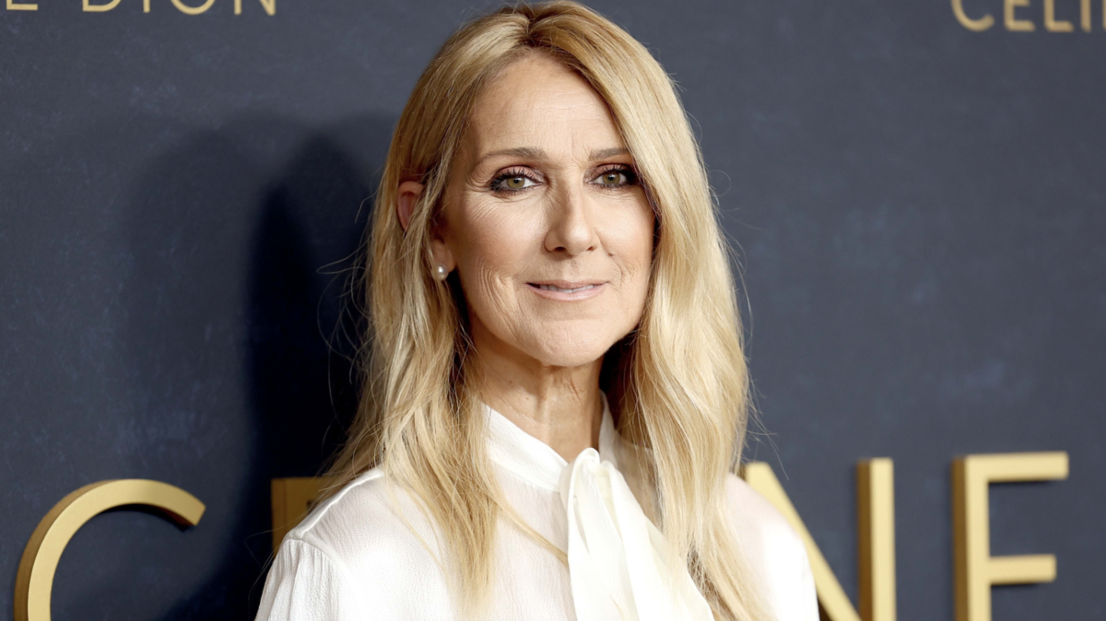 Celine Dion at an event, wearing a white, long-sleeved blouse with a tie at the neck, smiling at the camera