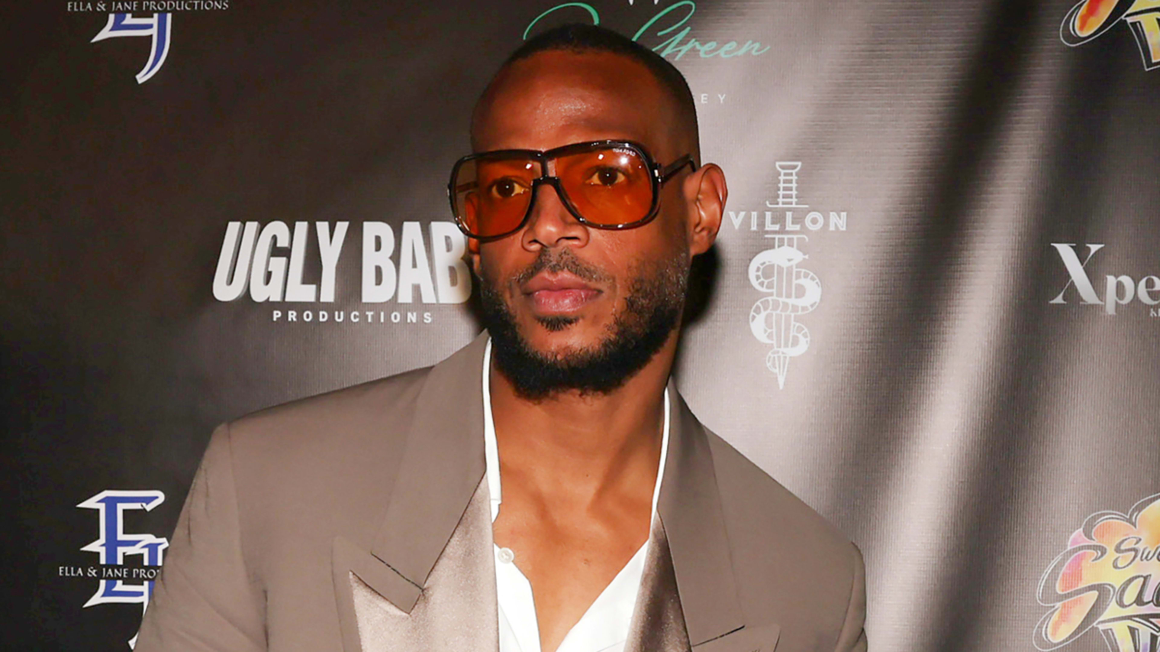 A man with a shaved head, sunglasses, and a trimmed beard wearing a tailored suit with a satin lapel stands at a media event backdrop