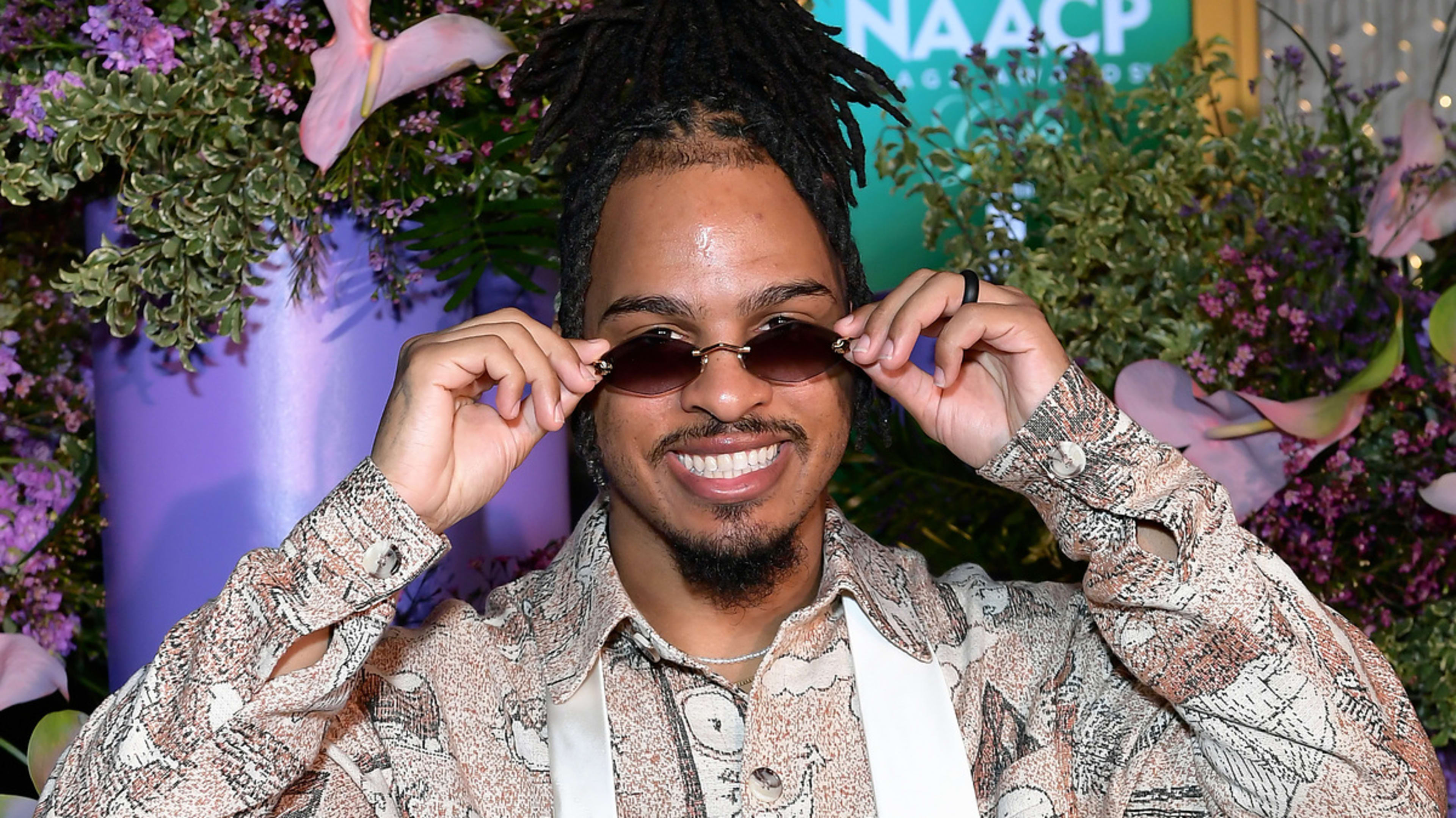 Man with dreadlocks, smiling while adjusting his sunglasses, in front of floral backdrop at the 55th NAACP event