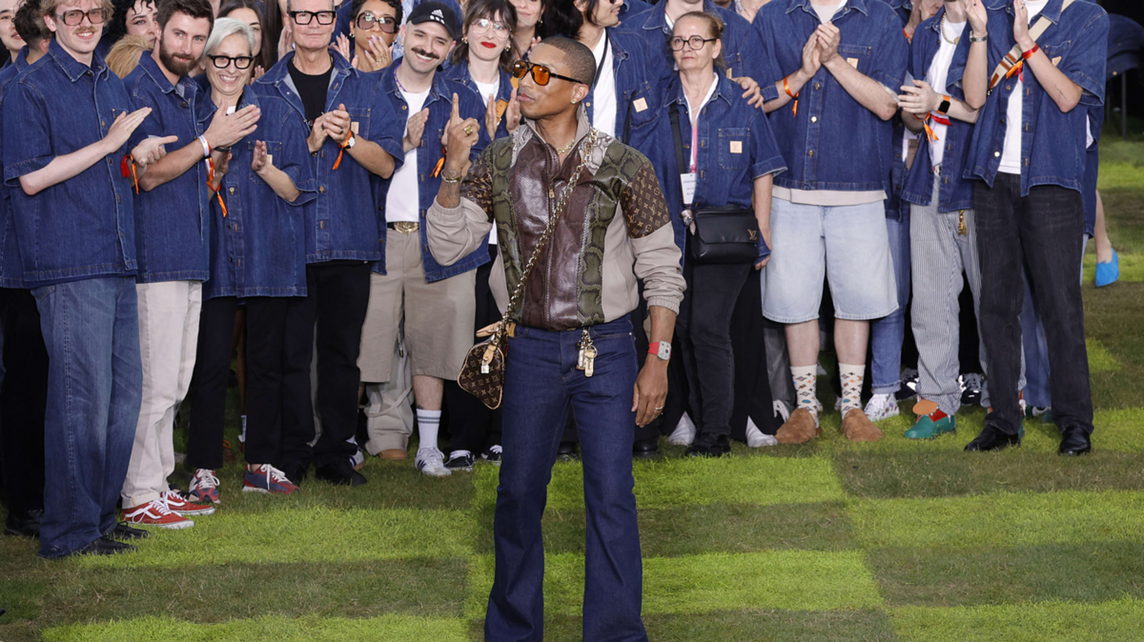 Pharrell Williams walks in front of a group of clapping individuals at a fashion event, wearing a stylish jacket and bell-bottom jeans