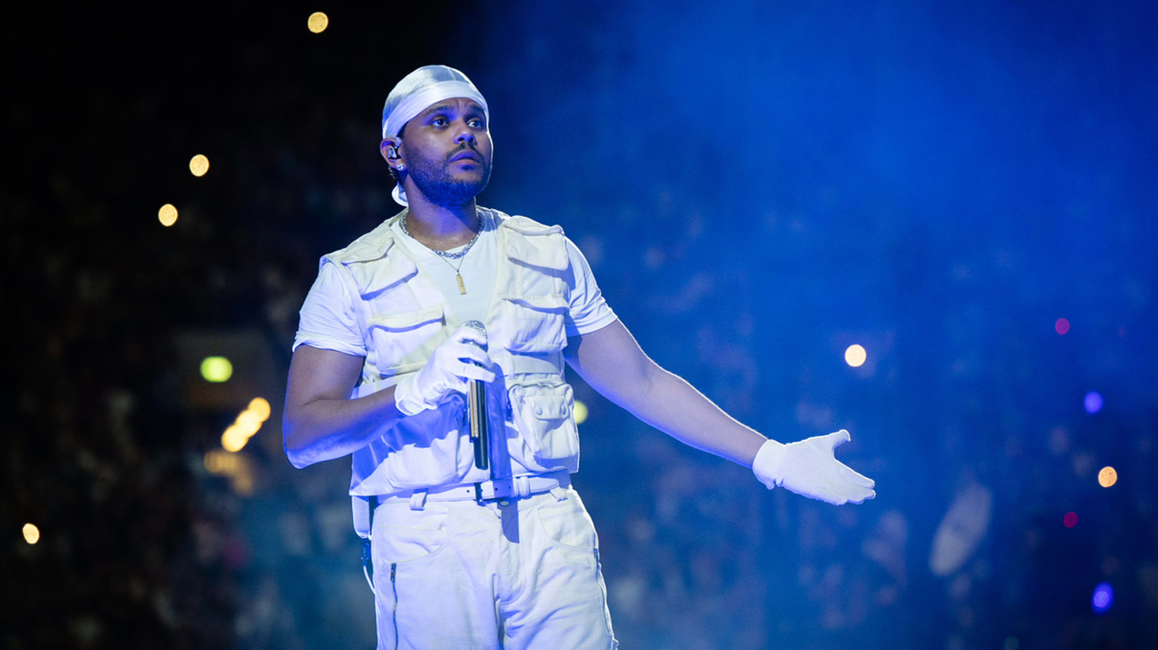 The Weeknd performs on stage wearing a white outfit, including a vest, gloves, and a durag, holding a microphone