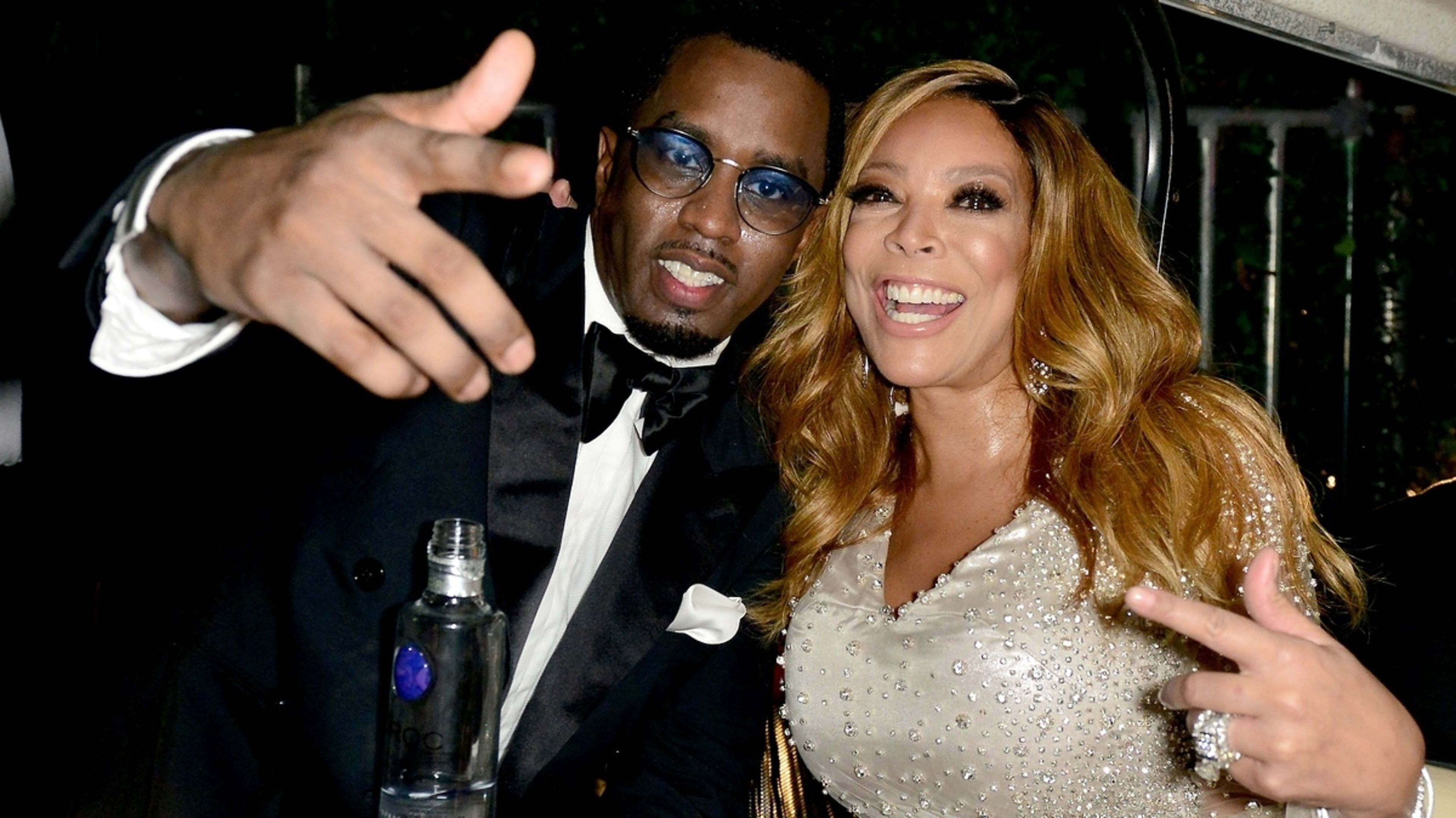 Sean “Diddy” Combs in a black tuxedo and Wendy Williams in a sequined dress smile and gesture at the camera